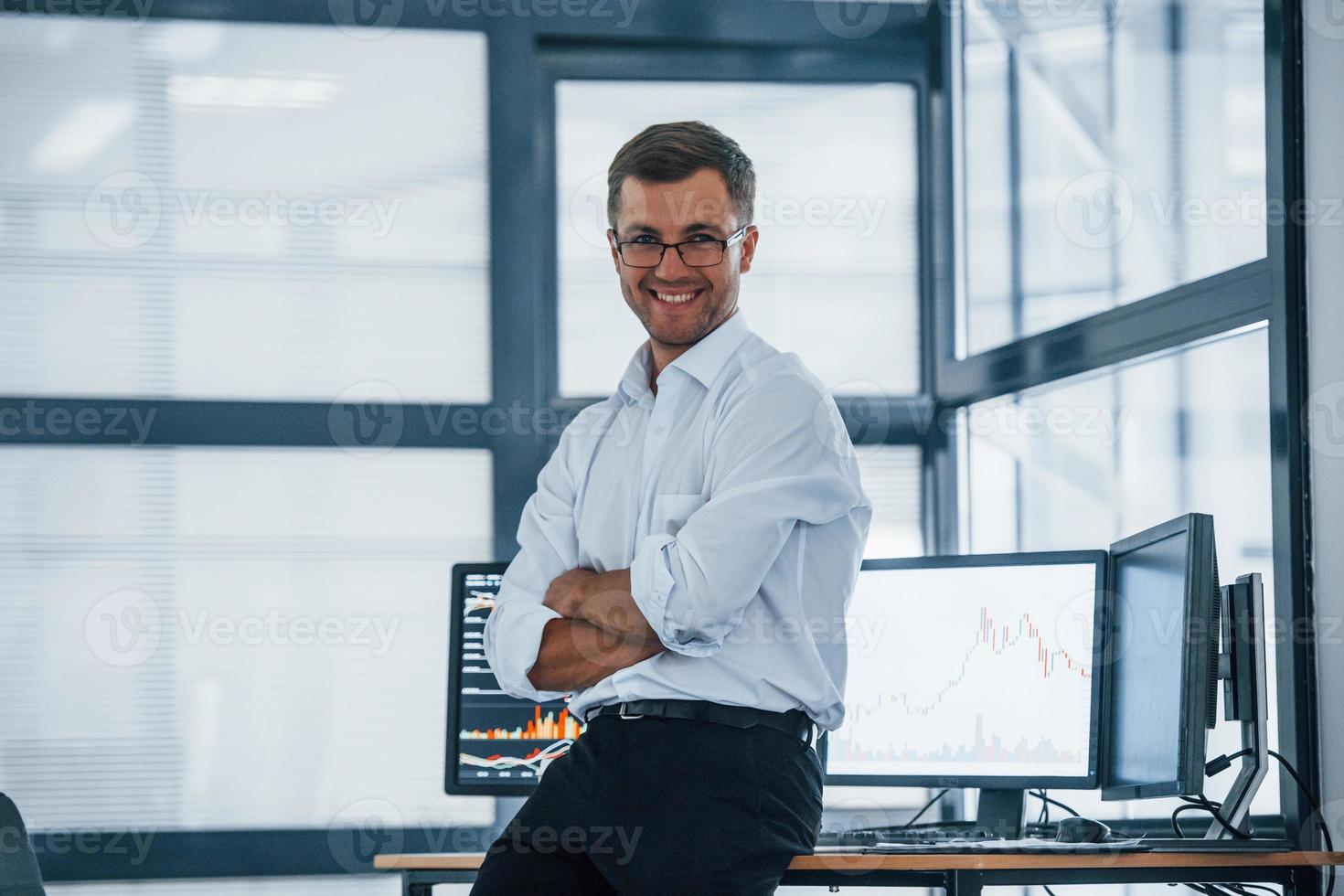 Happy employee. Young businessman in formal clothes is in office with multiple screens. Conception of exchange and money photo