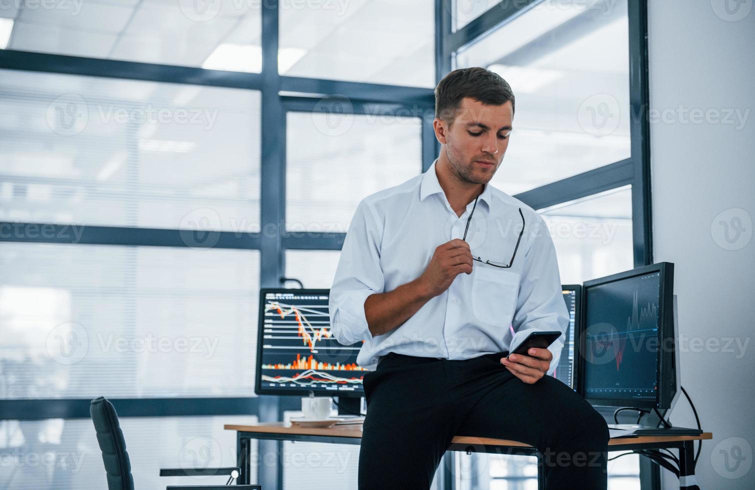 Sits on the table and having a break. With smartphone in hands. Young businessman in formal clothes is in office with multiple screens photo