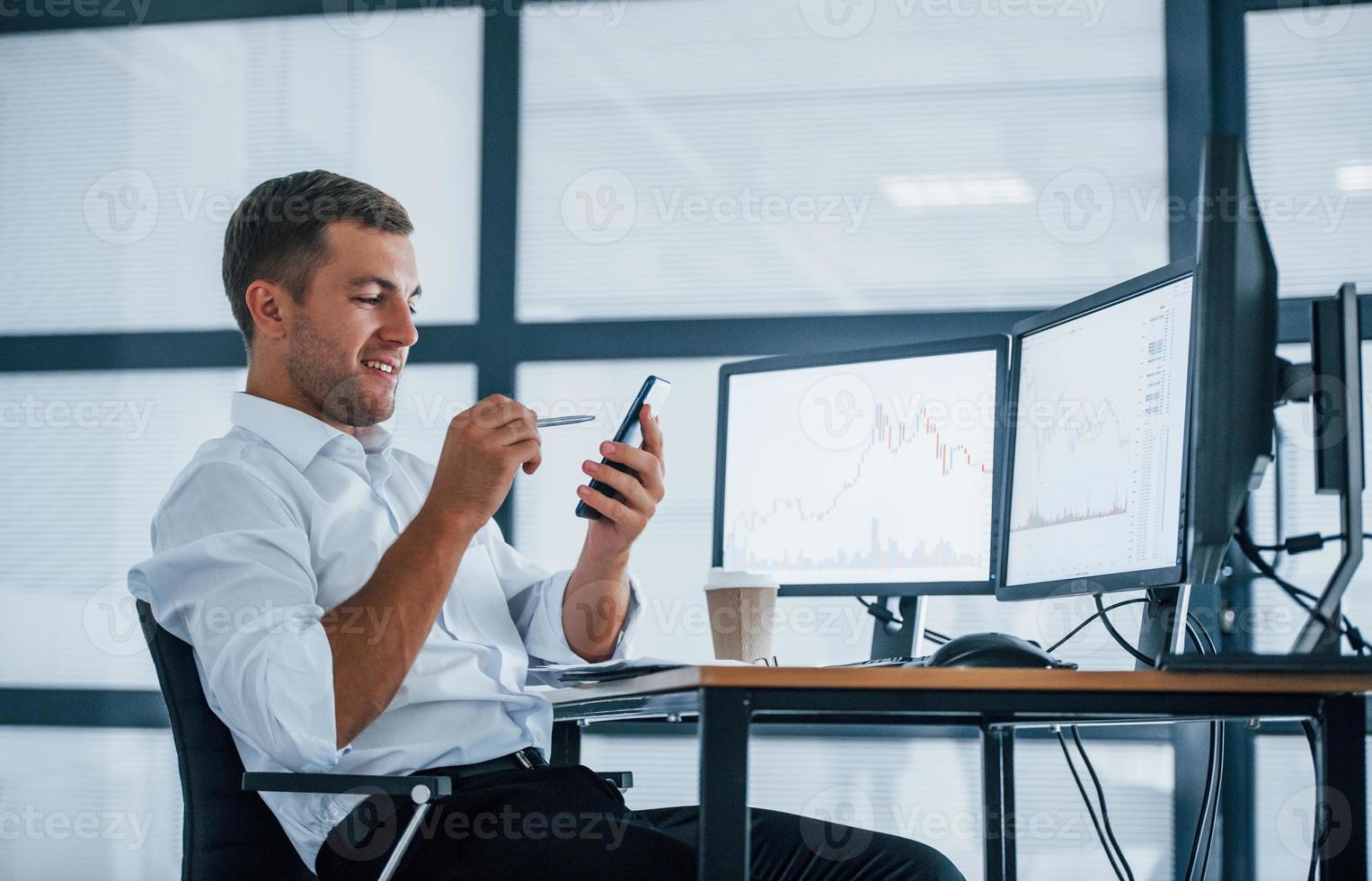 Young businessman in formal clothes is in office with multiple screens. Conception of exchange and money photo