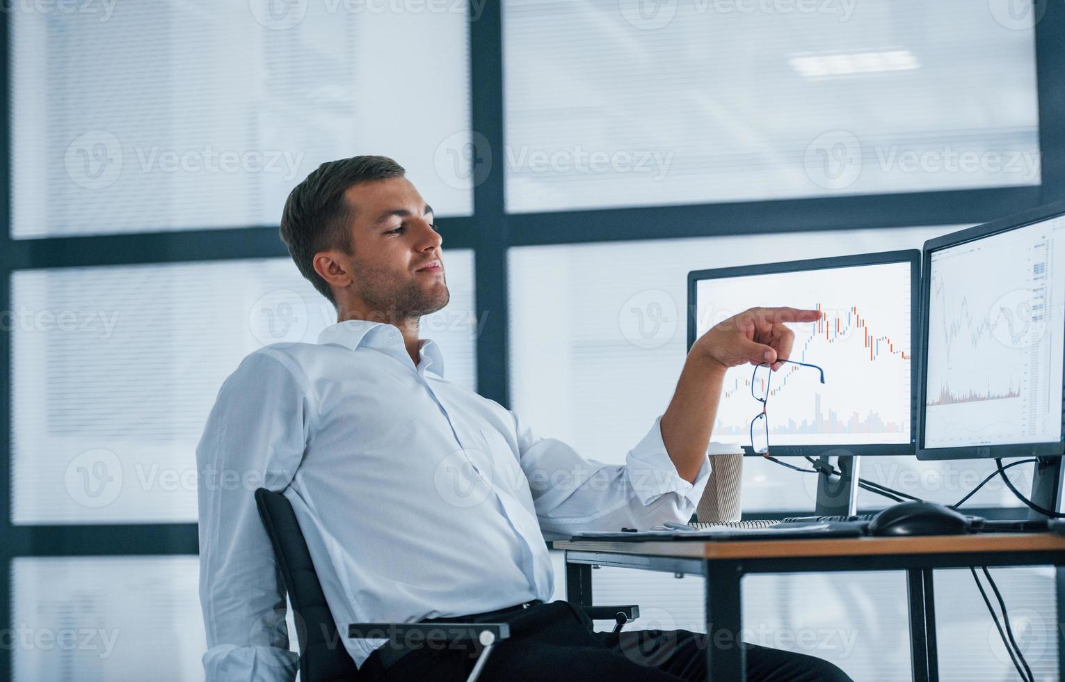 Feeling good. Conception of success. Young businessman in formal clothes is in office with multiple screens. Conception of exchange and money photo