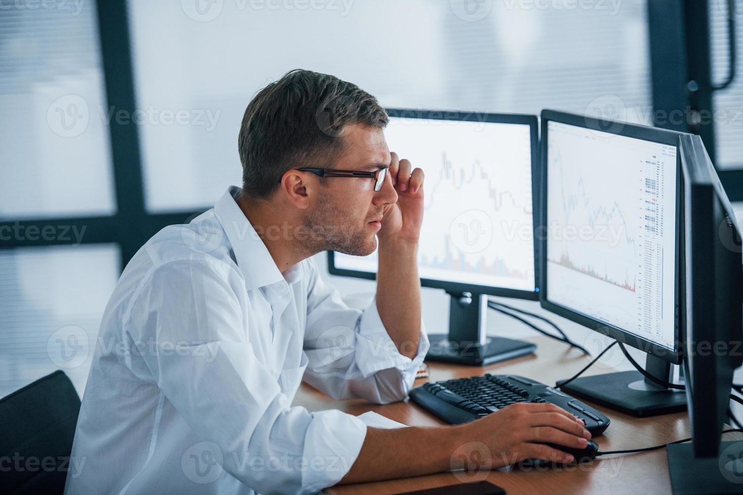 Young businessman in formal clothes is in office with multiple screens. Conception of exchange and money photo