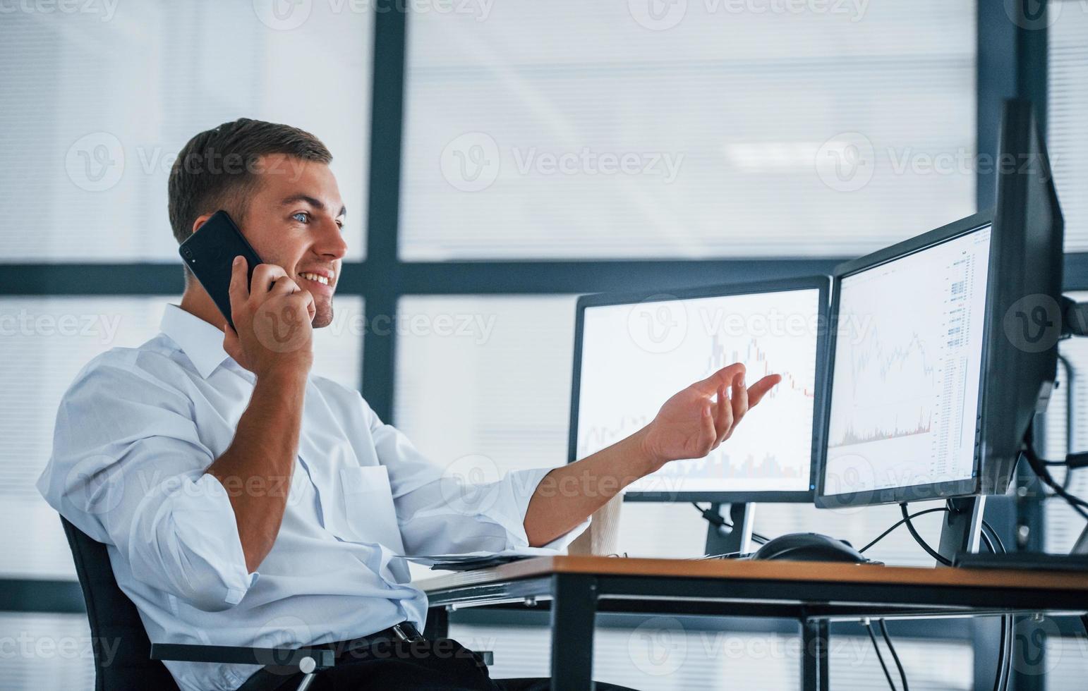 Having conversation by the phone. Young businessman in formal clothes is in office with multiple screens. Conception of exchange and money photo