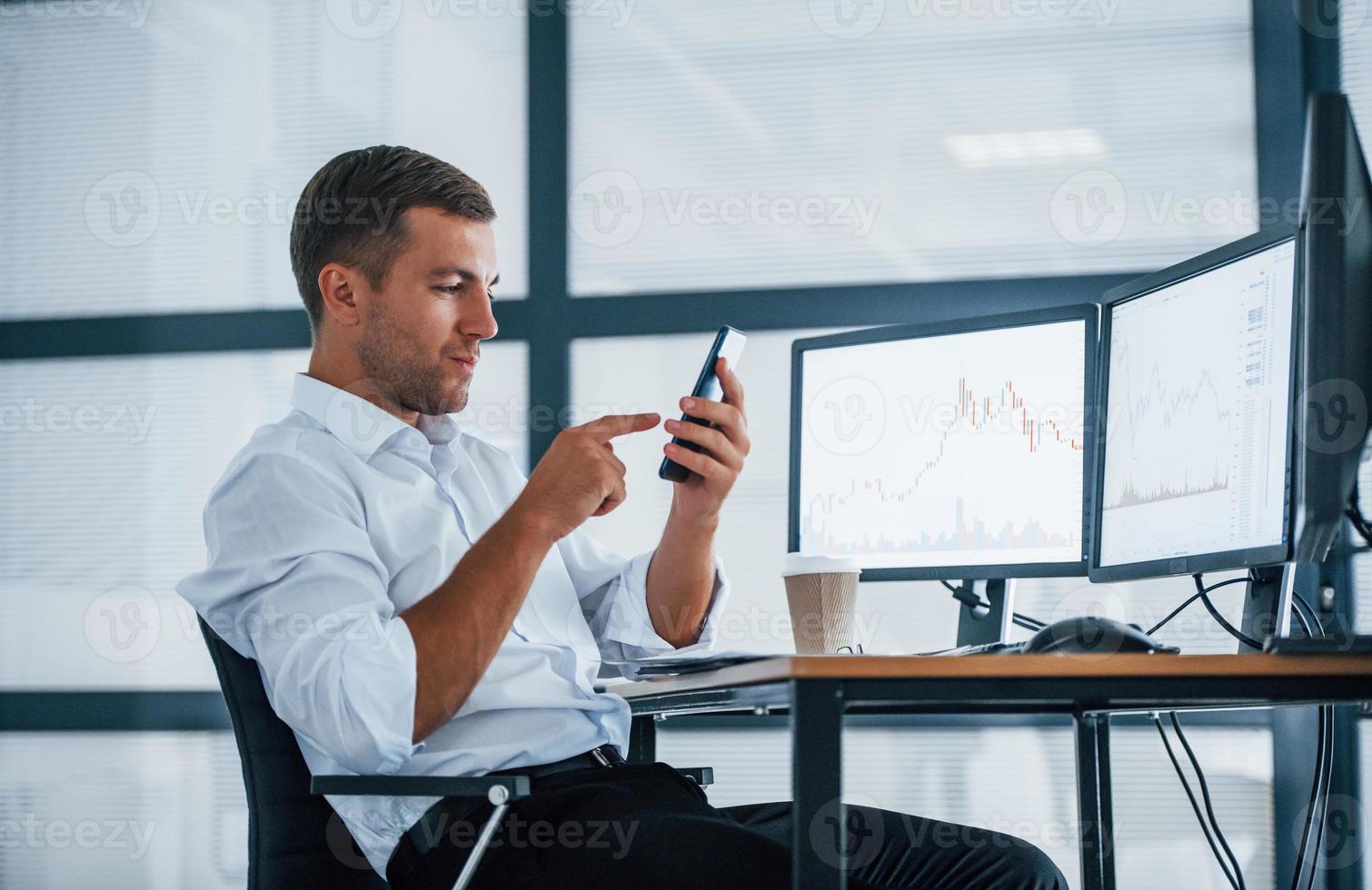 Young businessman in formal clothes is in office with multiple screens. Conception of exchange and money photo