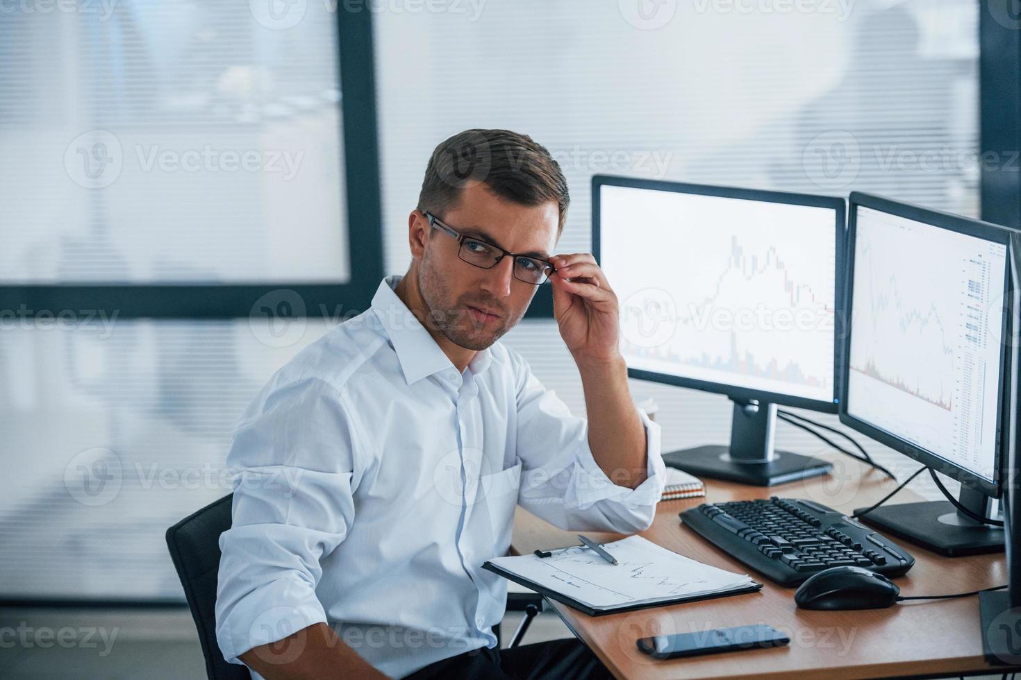 Looks at the camera. Young businessman in formal clothes is in office with multiple screens. Conception of exchange and money photo