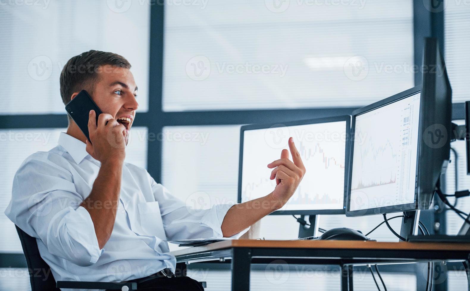 Having conversation by the phone. Young businessman in formal clothes is in office with multiple screens. Conception of exchange and money photo