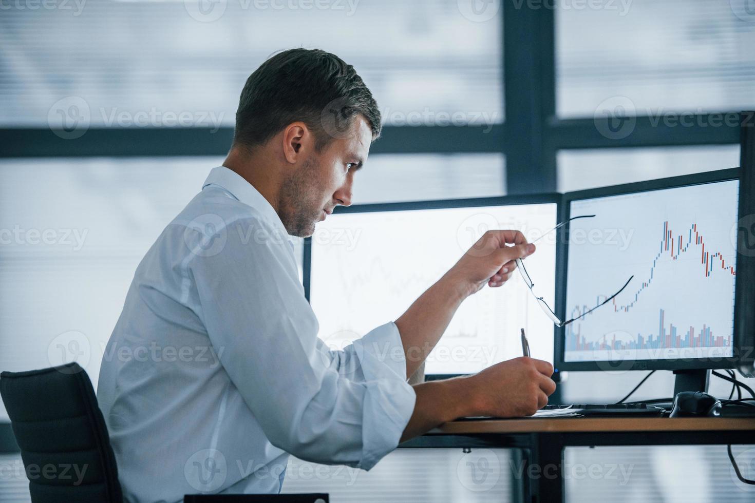 Side view. Young businessman in formal clothes is in office with multiple screens. Conception of exchange and money photo