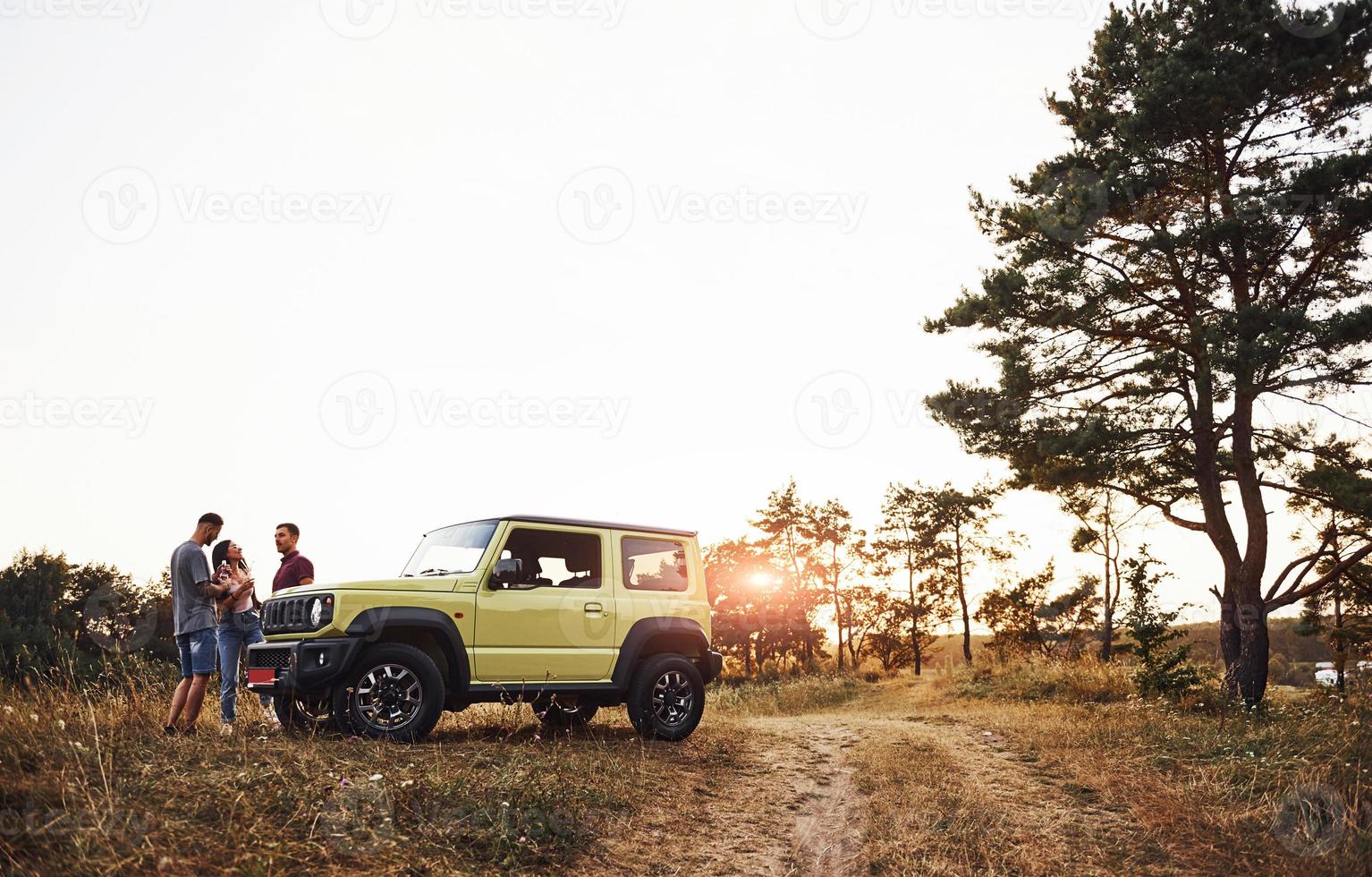 escena rural. grupo de amigos alegres que tengan un buen fin de semana en un día soleado cerca de su auto verde al aire libre foto