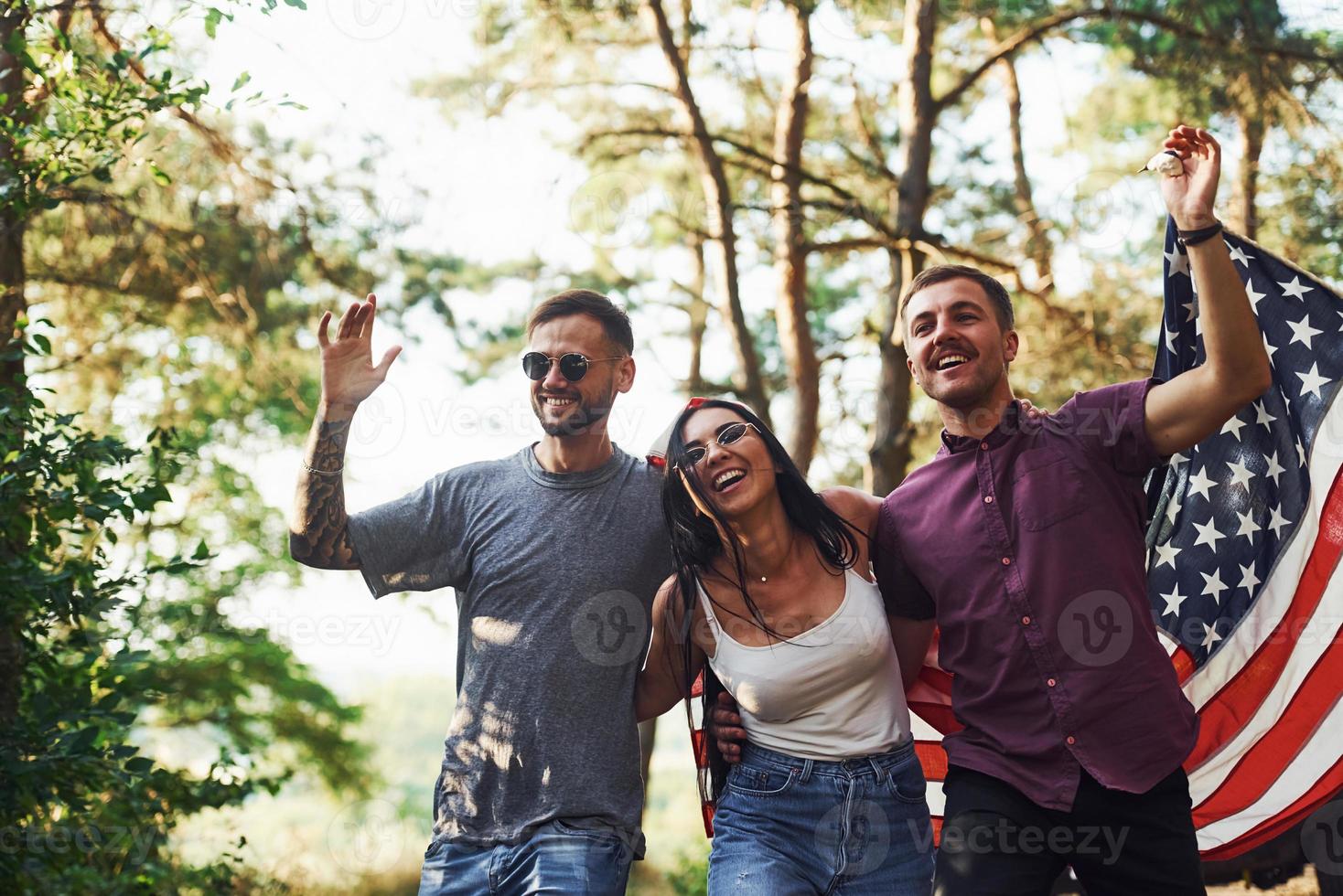 en el bosque. los amigos tienen un buen fin de semana al aire libre cerca de su auto verde con bandera de estados unidos foto