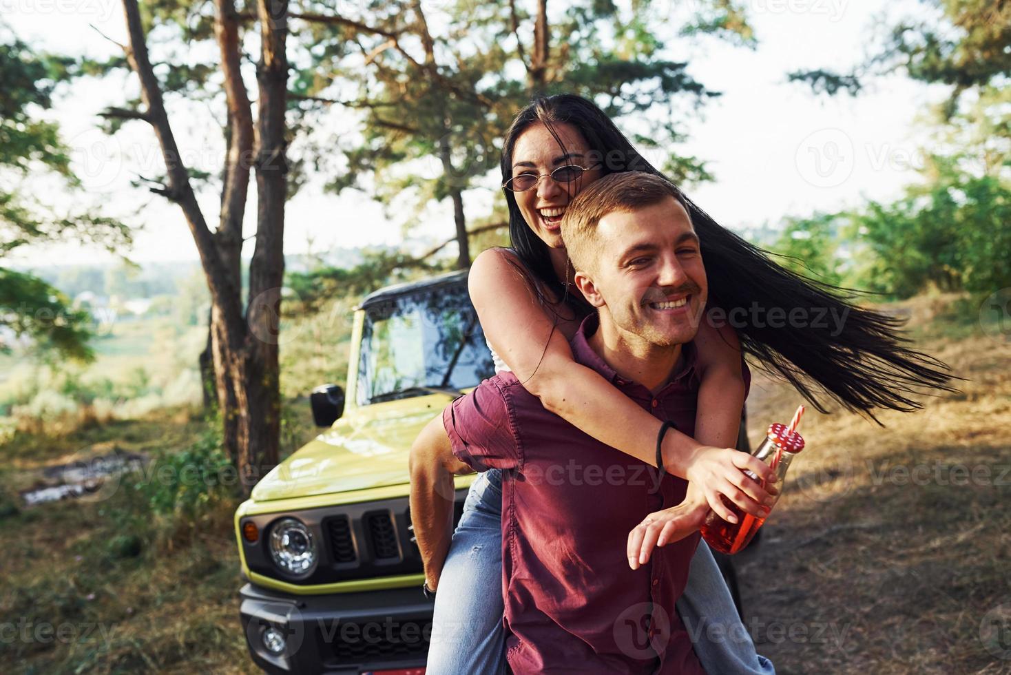 sonriendo y divirtiéndose. un par de jóvenes con alcohol se divierten en el bosque. jeep verde detrás foto