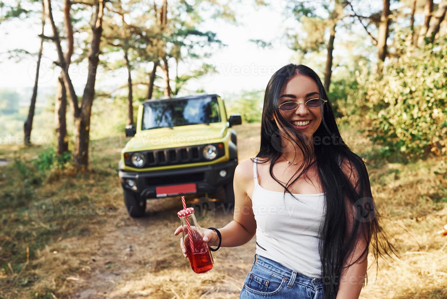 rubia con alcohol en la mano da un paseo por el bosque contra el jeep verde foto