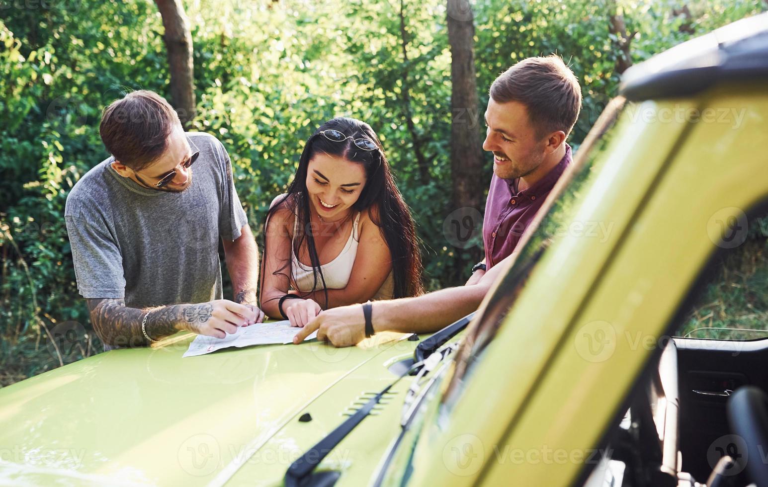 Young friends reading map that is on the hood of modern green jeep in the forest photo