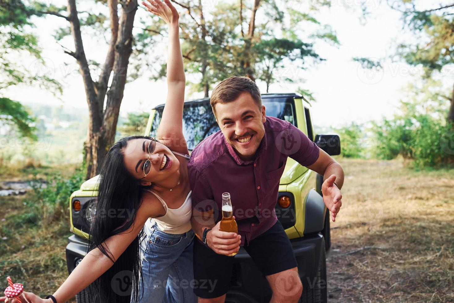 Smiling and having fun. Couple of young people with alcohol have fun in the forest. Green jeep behind photo