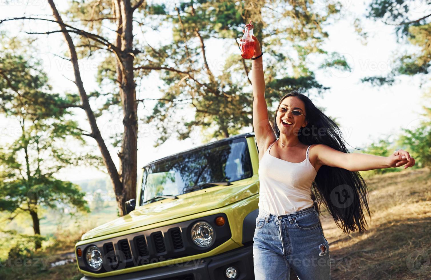 rubia con alcohol en la mano da un paseo por el bosque contra el jeep verde foto