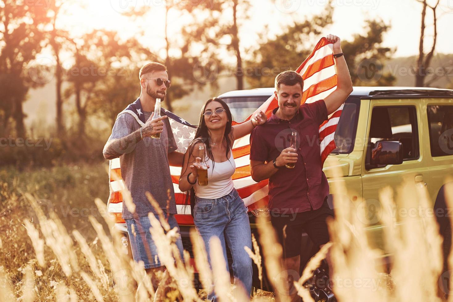 con alcohol en las manos. los amigos tienen un buen fin de semana al aire libre cerca de su auto verde con bandera de estados unidos foto