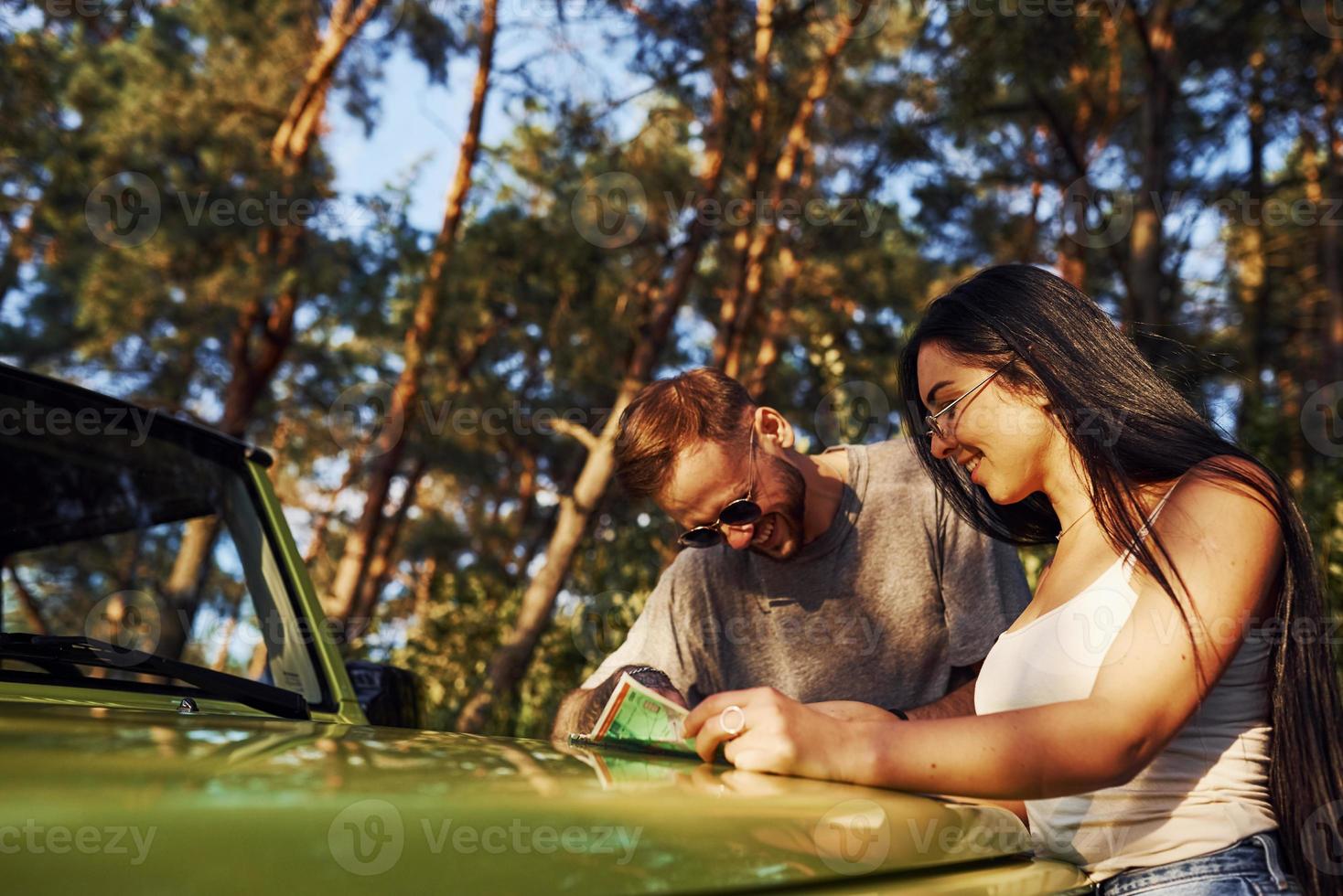 Young friends reading map that is on the hood of modern green jeep in the forest photo