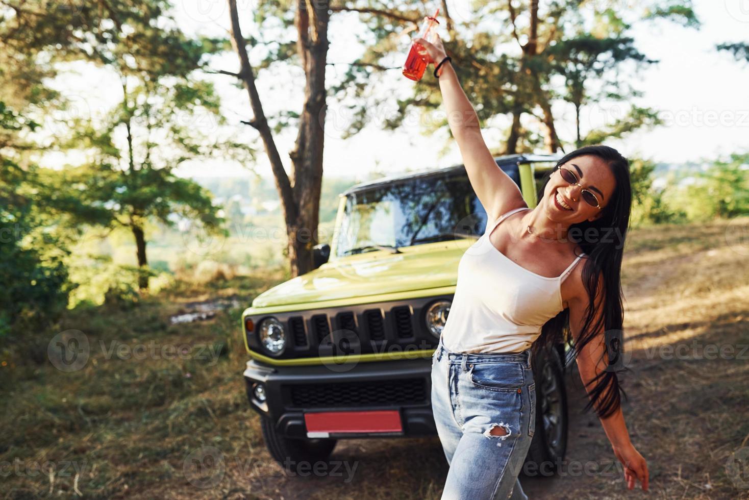 rubia con alcohol en la mano da un paseo por el bosque contra el jeep verde foto