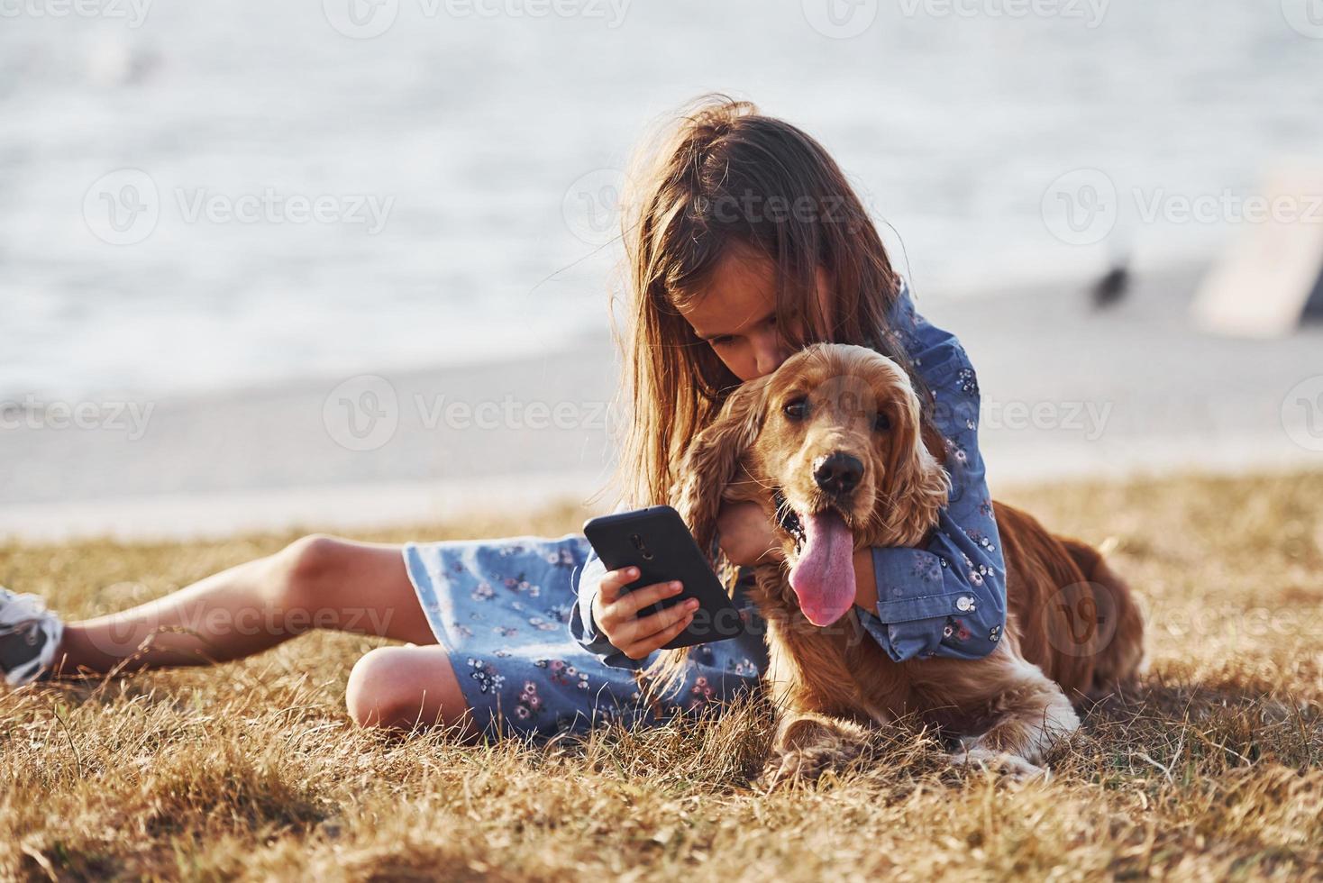 Lake behind. With phone in hands. Cute little girl have a walk with her dog outdoors at sunny day photo
