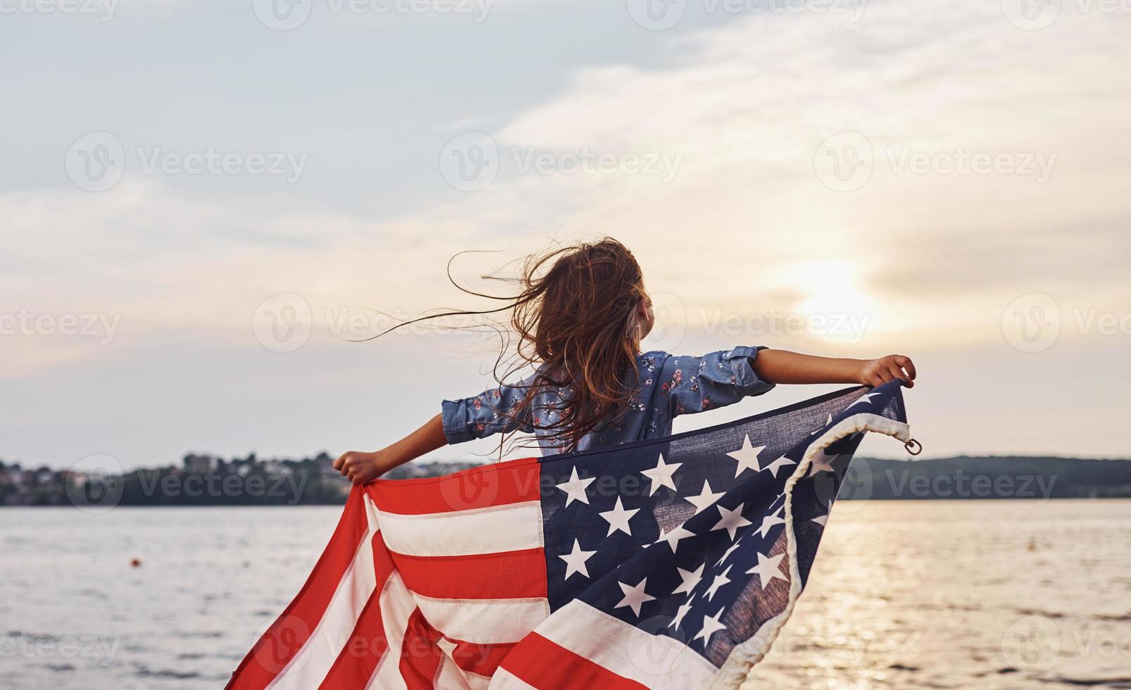 maravilloso atardecer. niña patriótica con bandera americana en las manos. contra el cielo nublado foto