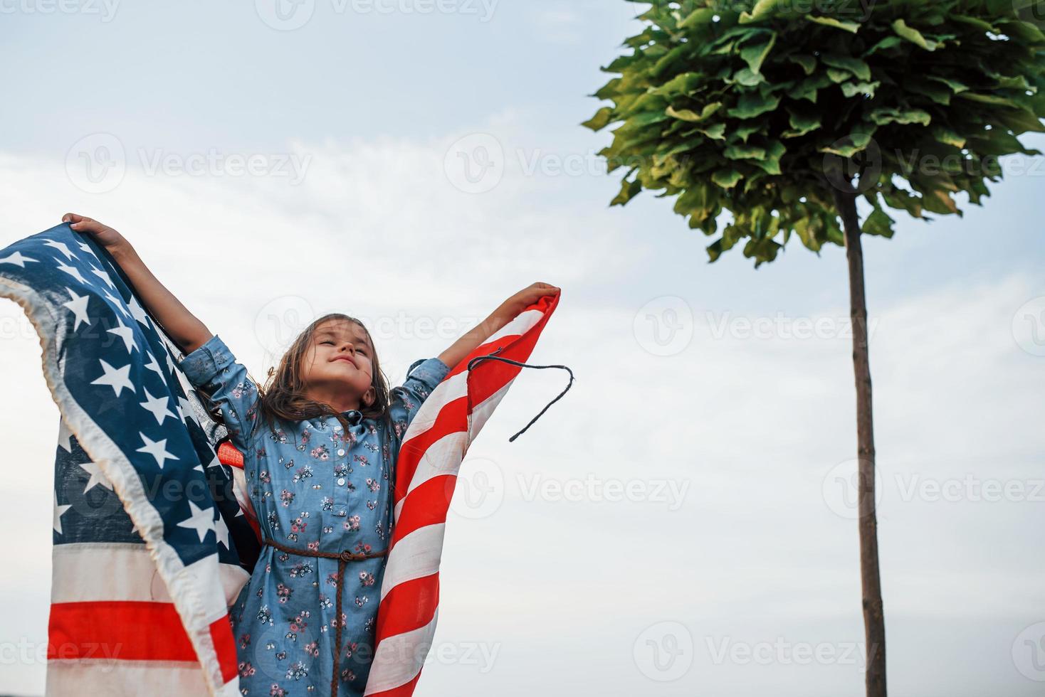 Beautiful green tree. Patriotic female kid with American Flag in hands. Against cloudy sky photo