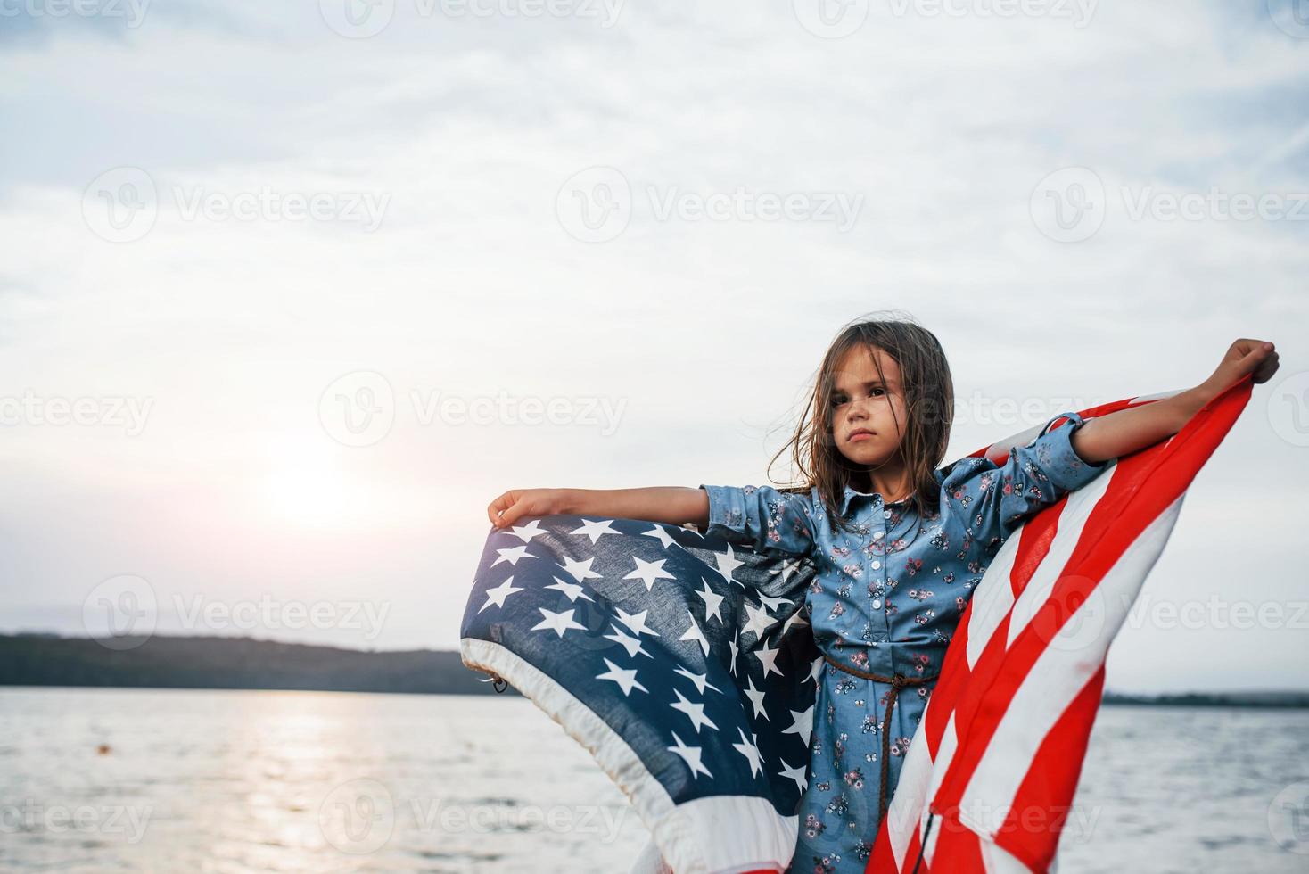 Patriotic female kid with American Flag in hands. Against cloudy sky photo