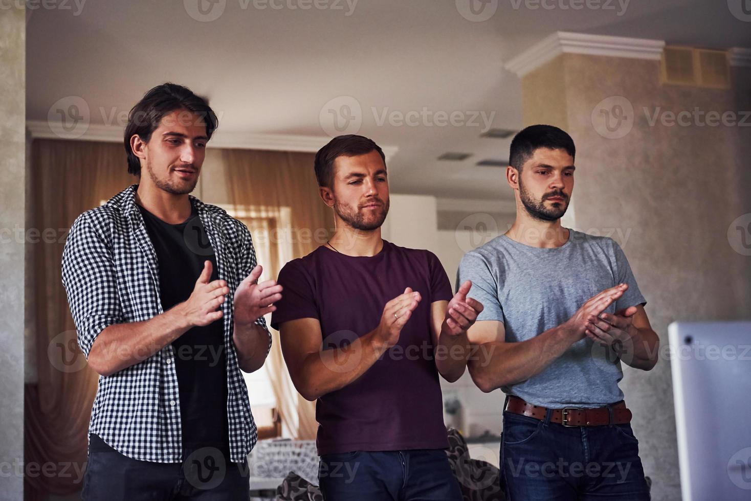 tres amigos se paran en la sala de estar y aplauden contra la televisión foto