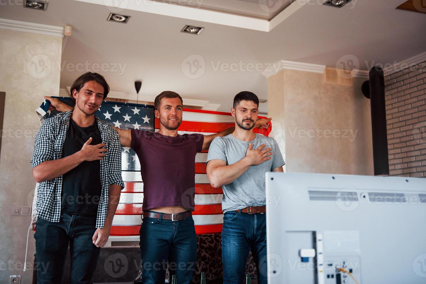 Adult patriots standing against TV with USA flag and hands to the hearts. Hymn is playing photo