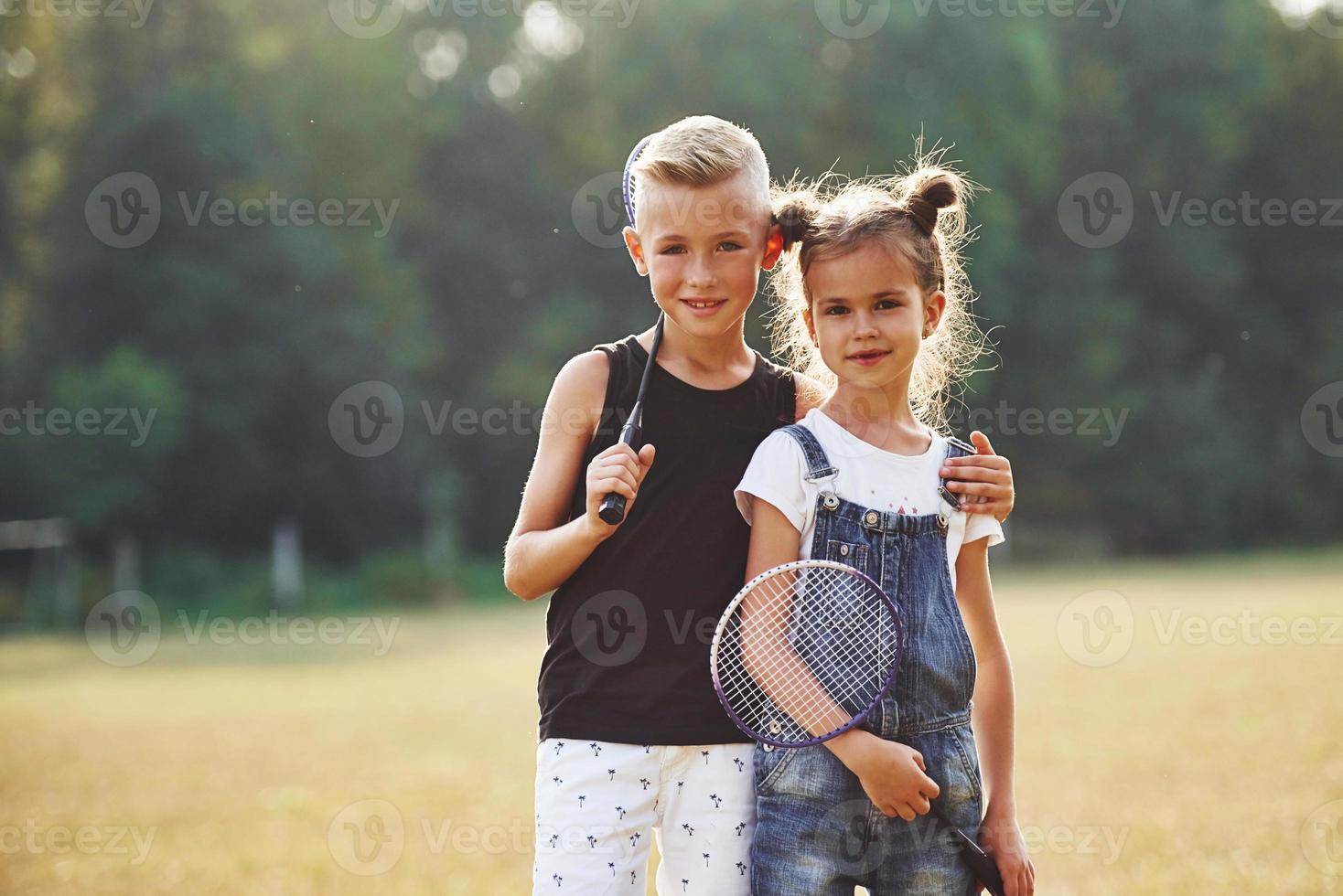 Portrait of boy and girl that standing in the field at sunny day with tennis rackets in hands photo