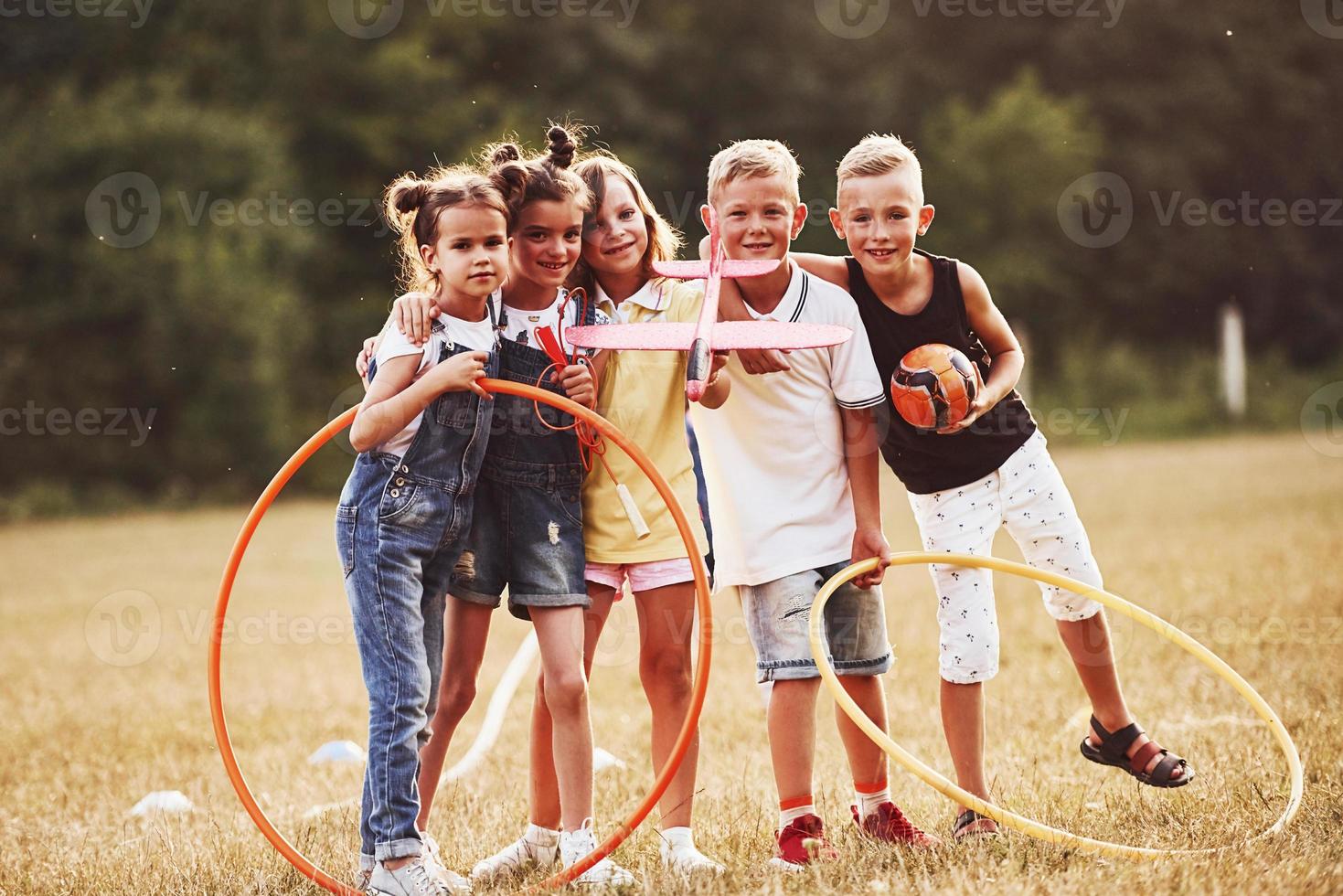 niños pequeños con pelota de fútbol, juguete de avión y círculos de fitness se paran juntos en el campo en un día soleado foto