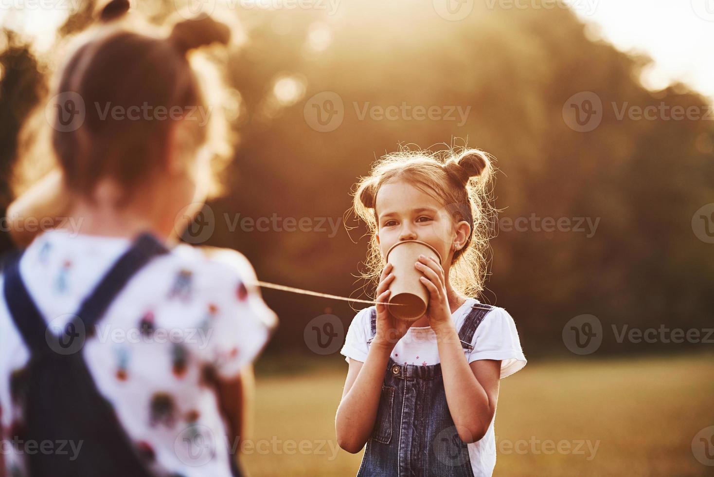 Two female kids stands in the field and talking by using string can phone photo