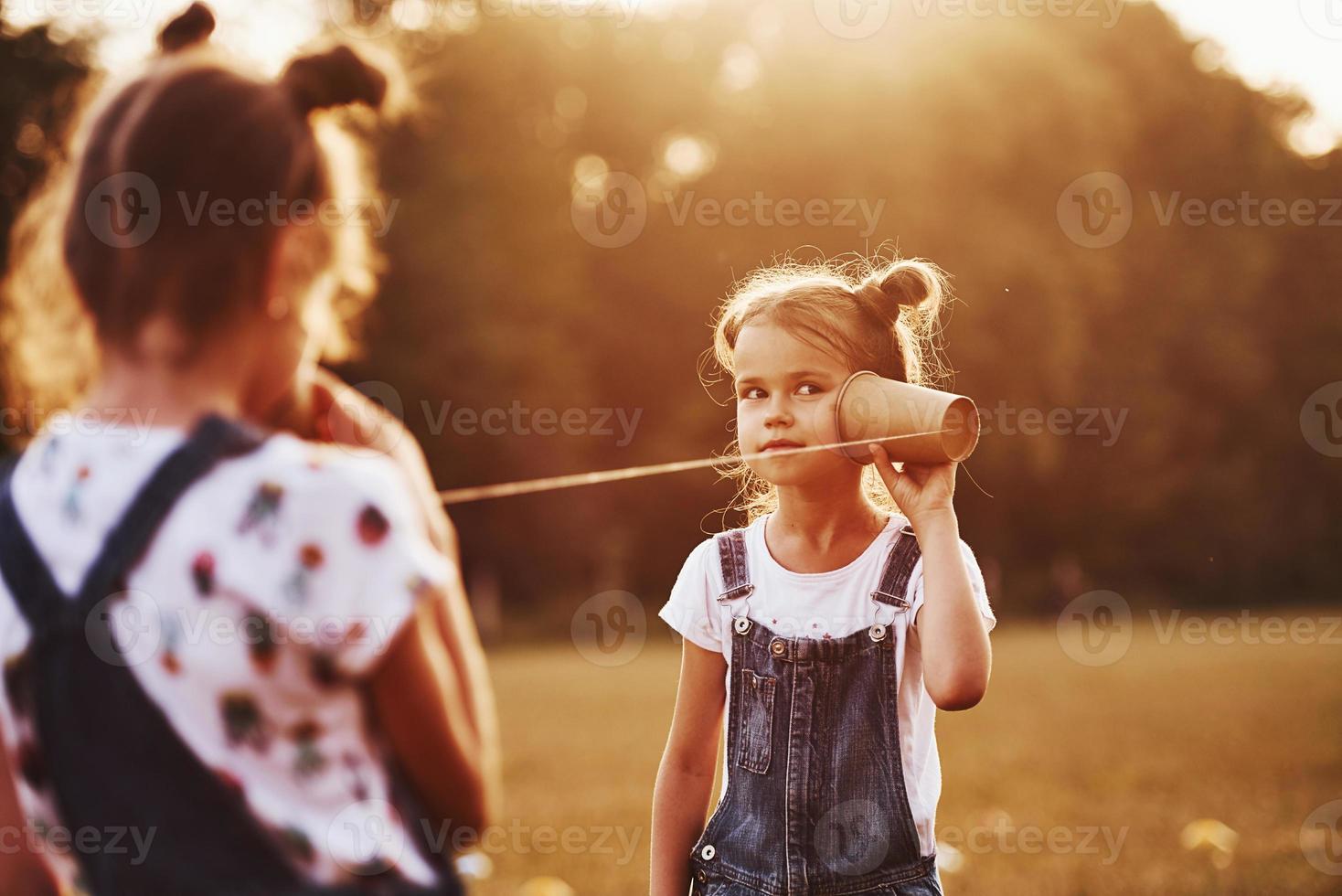 Two female kids stands in the field and talking by using string can phone photo