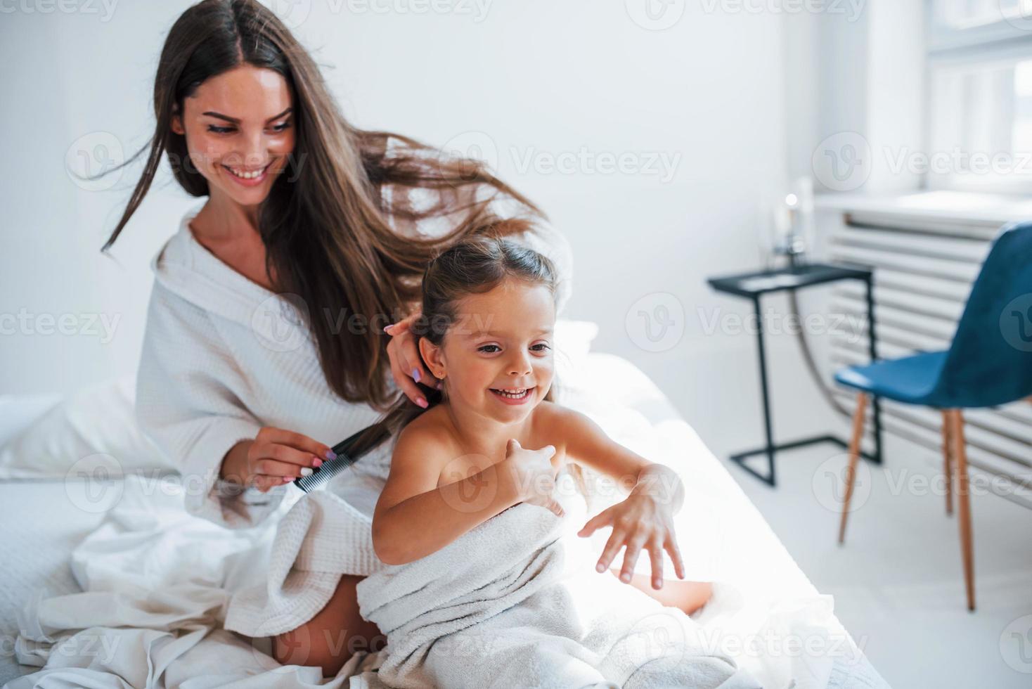 hacer peinado usando peine. la joven madre con su hija tiene un día de belleza en el interior de la habitación blanca foto