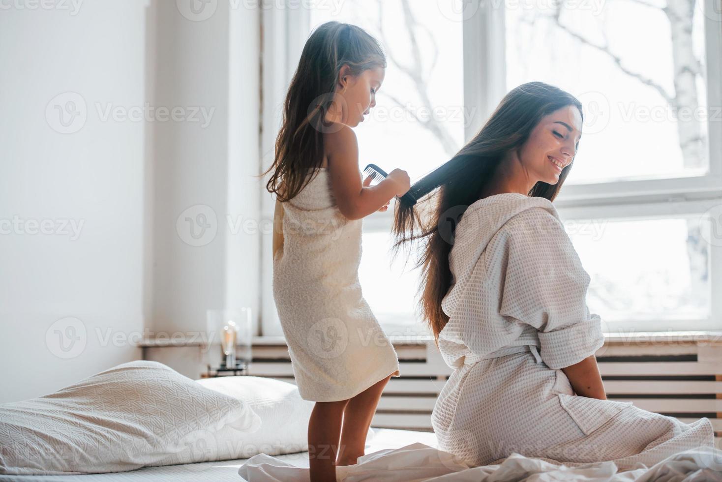 ayudando a hacer el peinado. la joven madre con su hija tiene un día de belleza en el interior de la habitación blanca foto