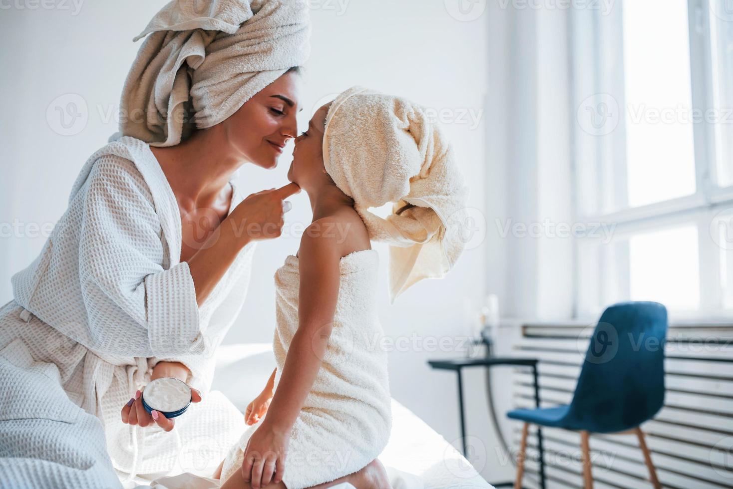 Using cream to clear skin. Young mother with her daugher have beauty day indoors in white room photo