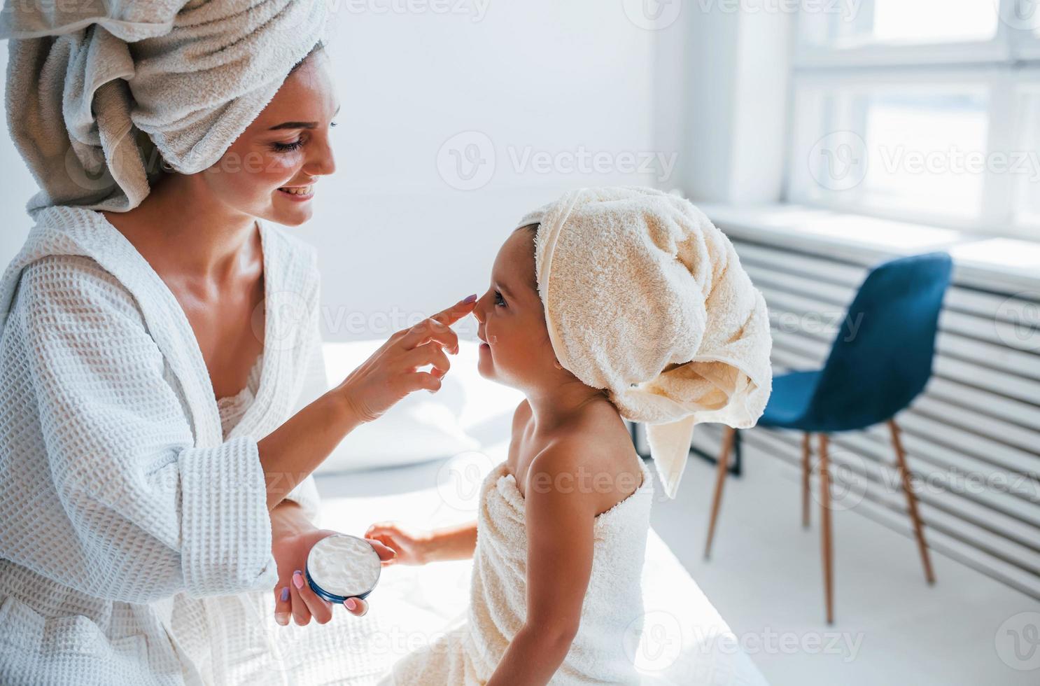 usando crema para aclarar la piel. la joven madre con su hija tiene un día de belleza en el interior de la habitación blanca foto