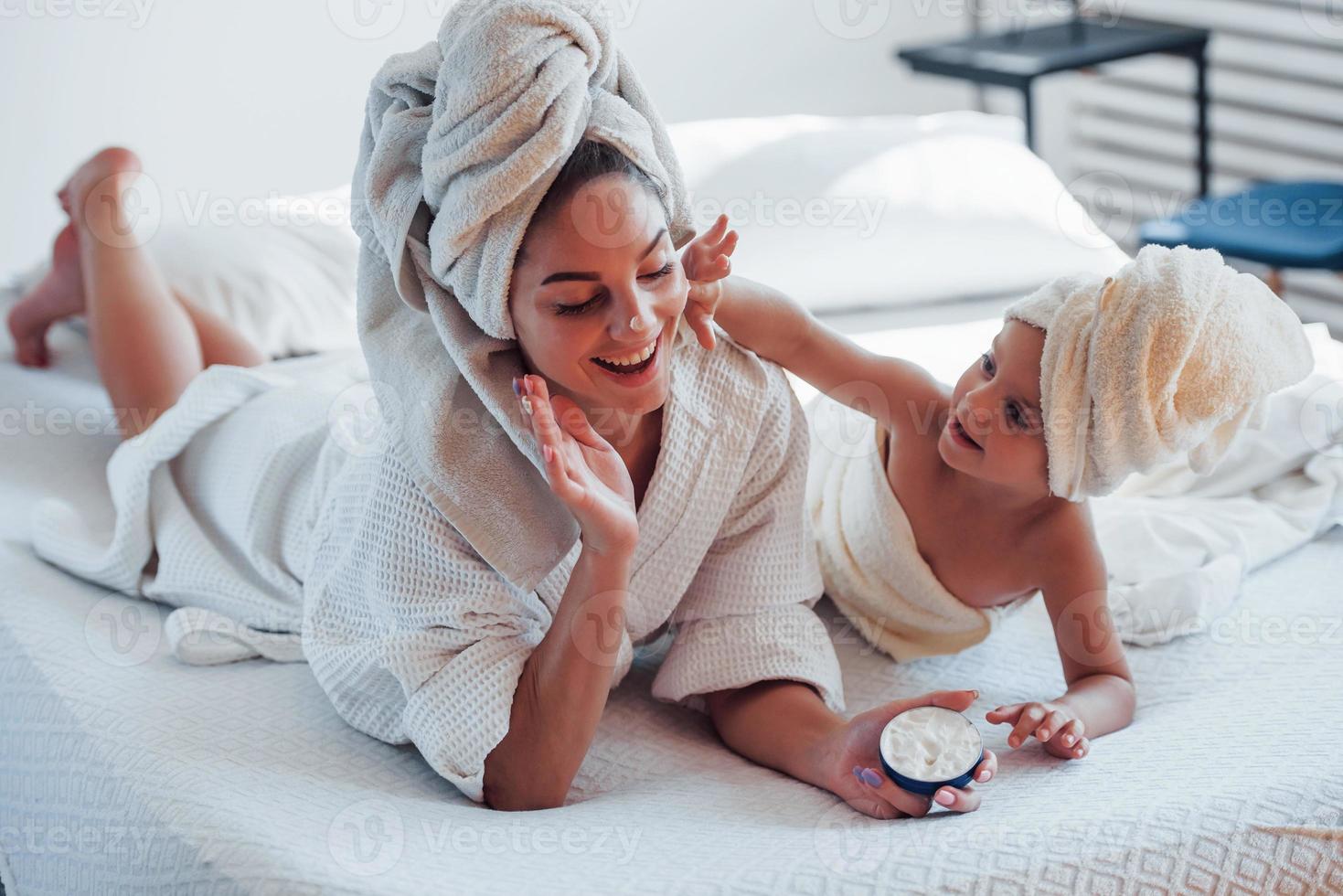 usando crema la joven madre con su hija tiene un día de belleza en el interior de la habitación blanca foto