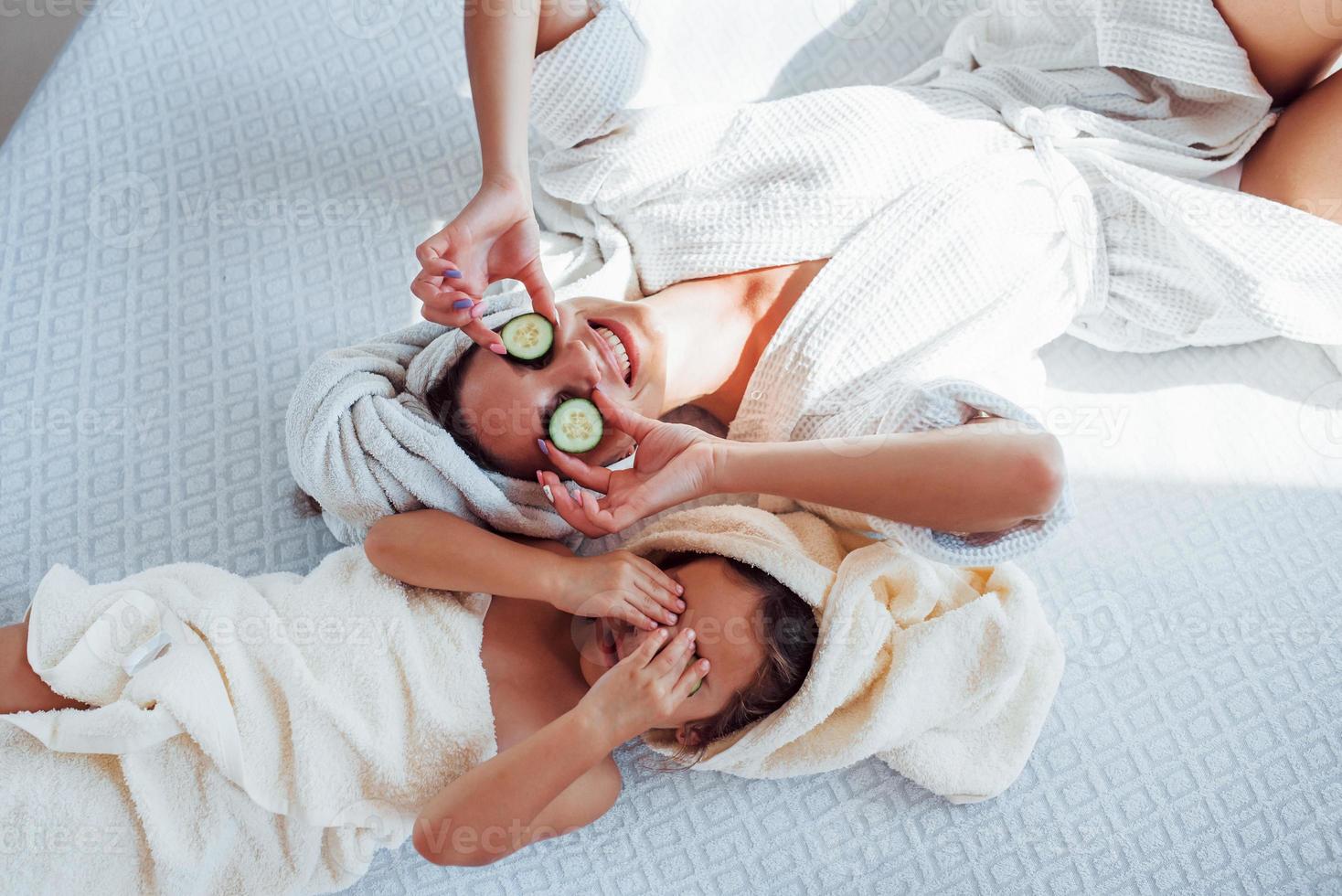 Young positive mother and daughter with towel on head lying on the bed together with cucumber photo