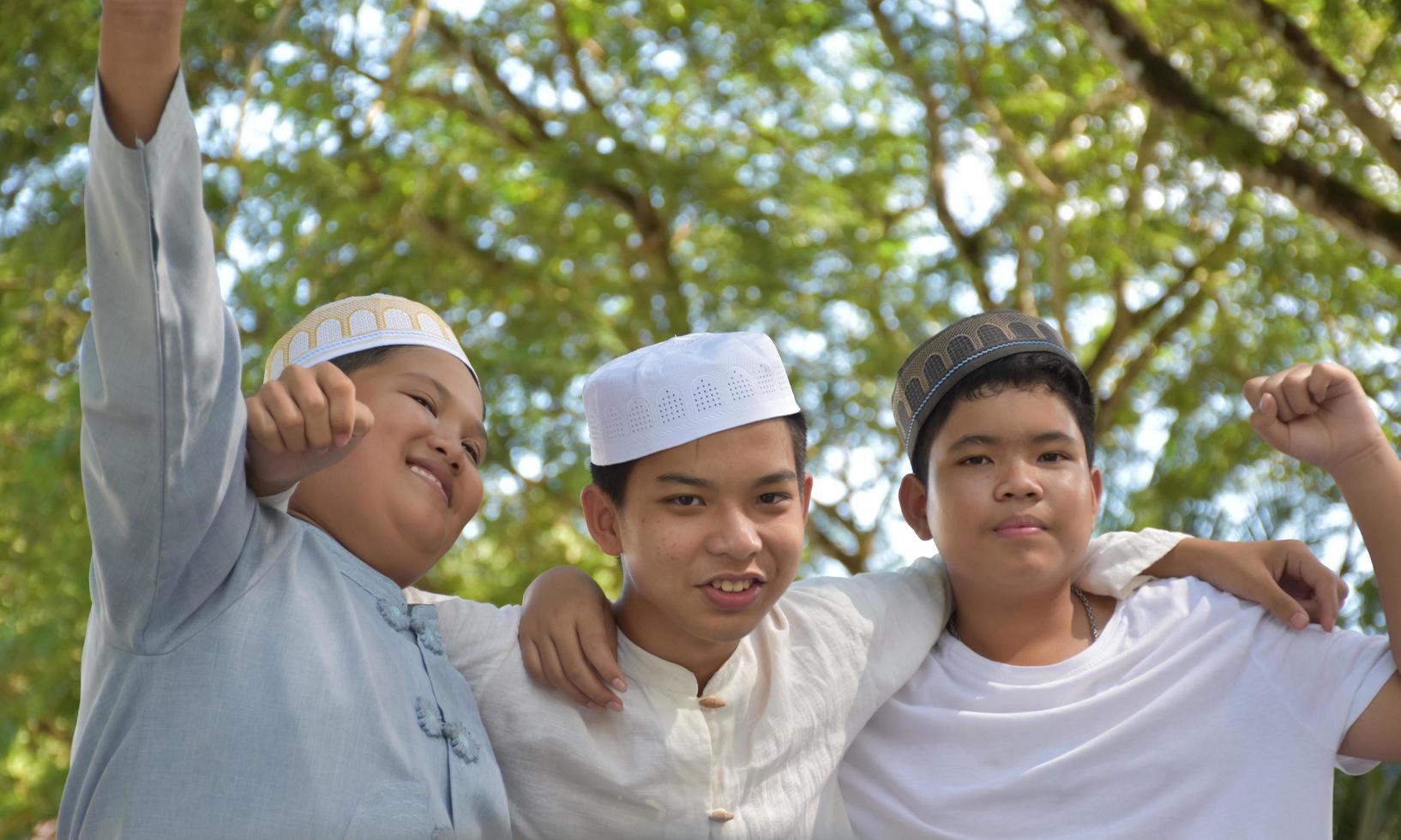 Young asian muslim boys raised hands, smilling and hugging each other to present happiness under the trees in the park, soft and selective focus. photo