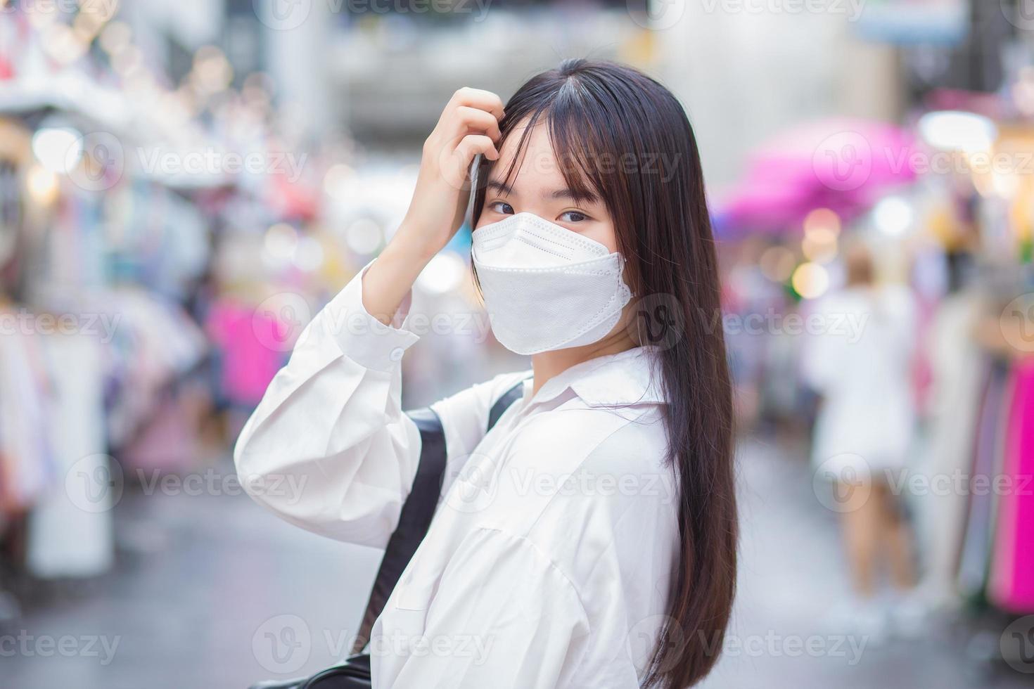 Linda joven estudiante asiática que usa camisa blanca y mascarilla para prevenir enfermedades de pie y sonriendo mientras está al aire libre en la ciudad con la calle con tiendas en el fondo. foto