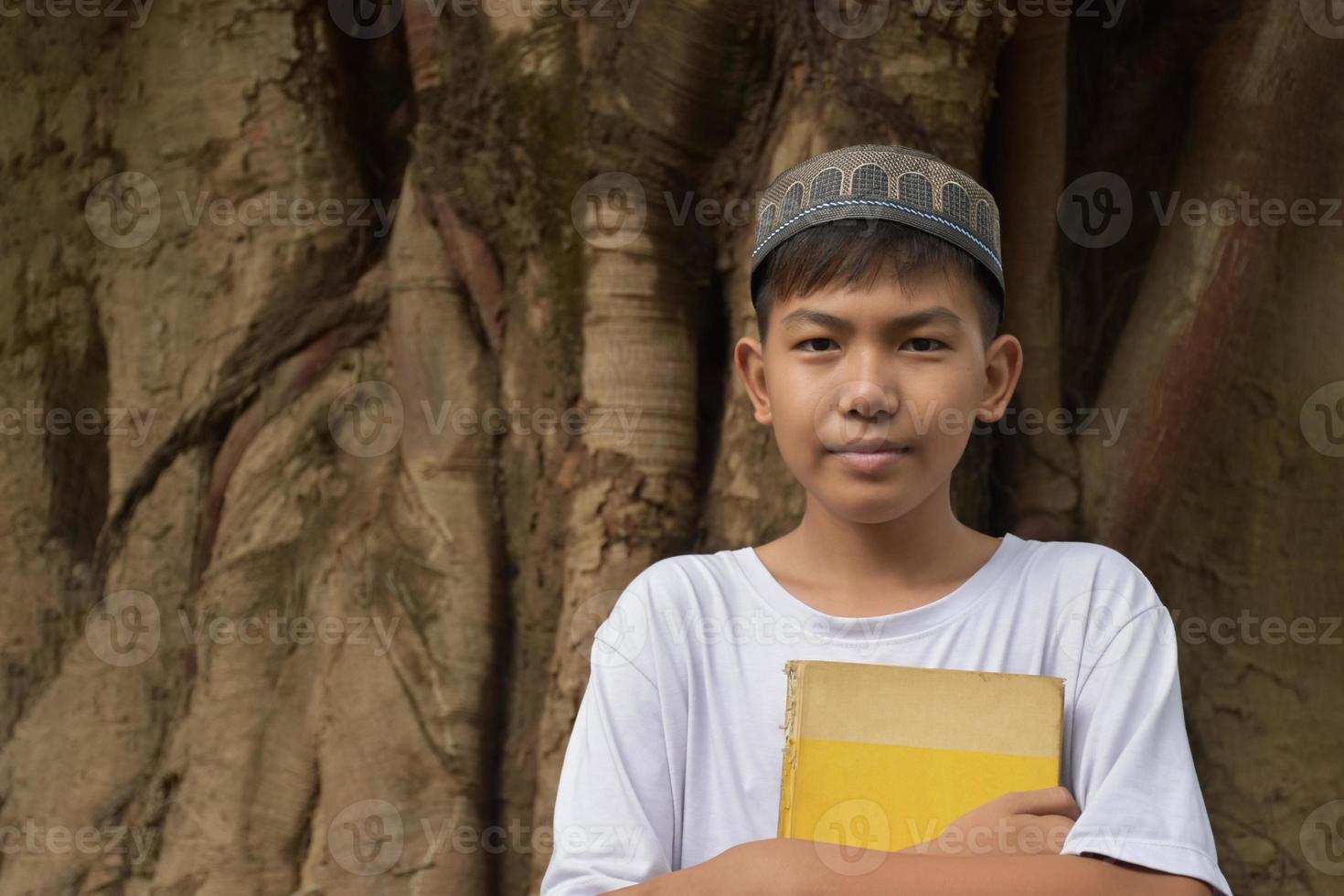 retrata a un niño musulmán o islámico asiático parado solo frente al gran árbol y sosteniendo un libro en el pecho, enfoque suave y selectivo. foto