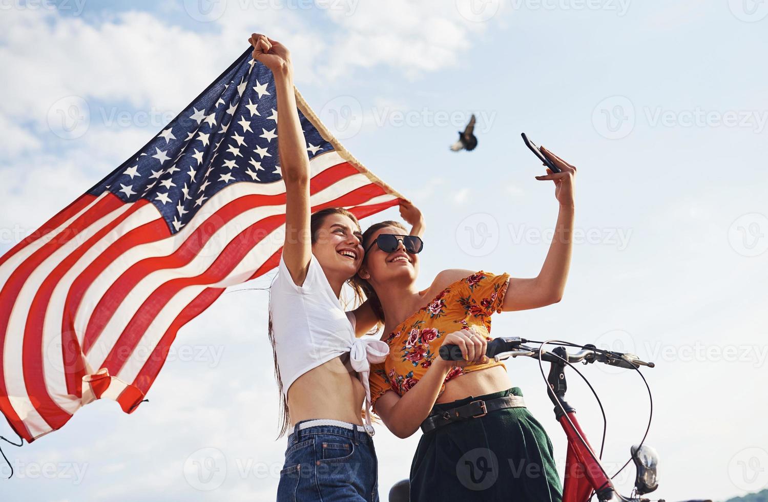 Against cloudy sky. Two patriotic cheerful woman with bike and USA flag in hands makes selfie photo