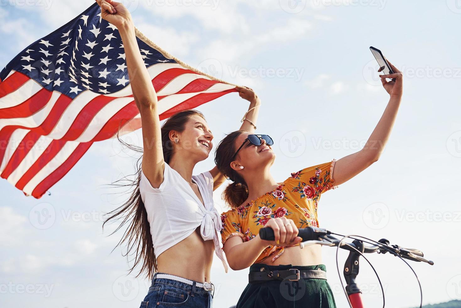 Against cloudy sky. Two patriotic cheerful woman with bike and USA flag in hands makes selfie photo