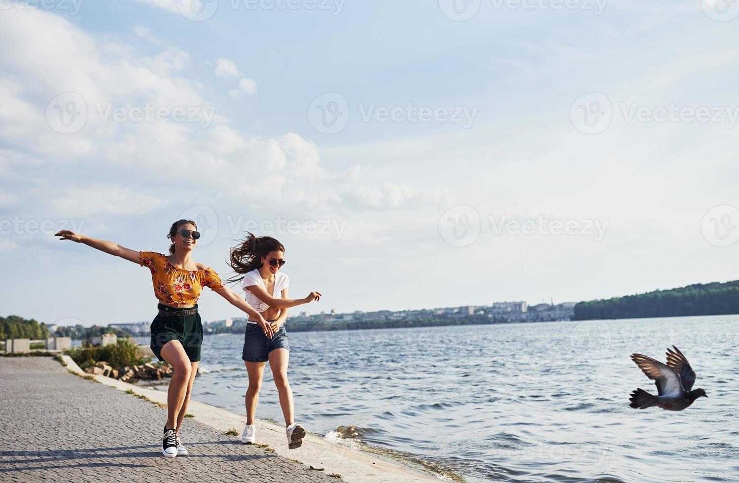 Bird flying in the air. Two female friends runs and have fun at beach near the lake at sunny daytime photo