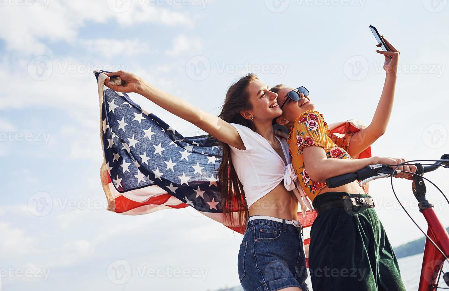 Two patriotic cheerful women with bike and USA flag in hands makes selfie photo
