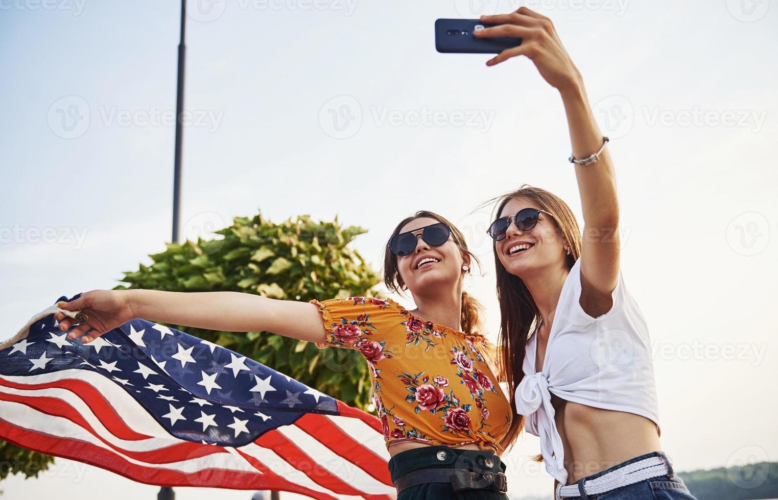Green tree at background. Two patriotic cheerful women with USA flag in hands making selfie outdoors in park photo