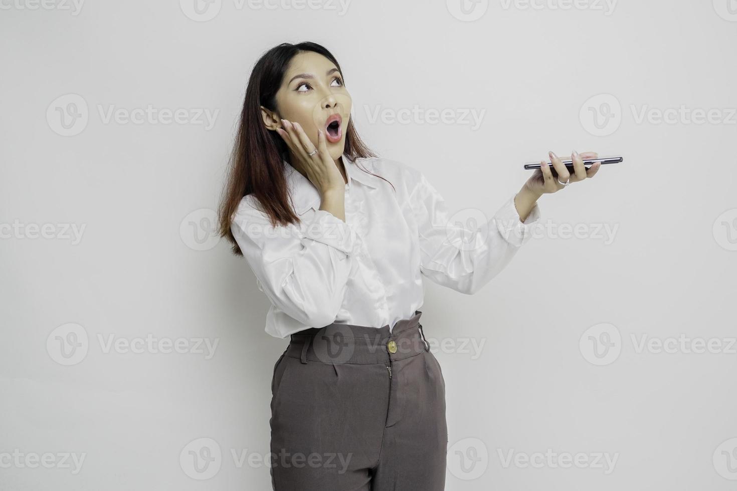 Shocked Asian woman wearing white shirt and holding her phone, isolated by white background photo