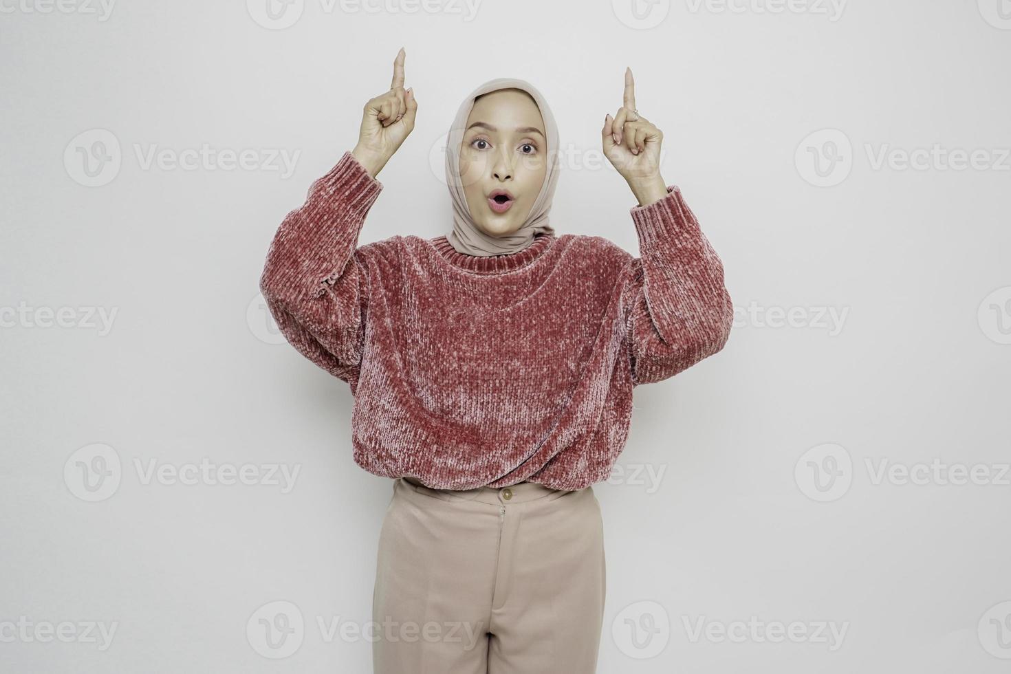 Shocked Asian Muslim woman wearing pink sweater and hijab pointing at the copy space on top of her, isolated by white background photo