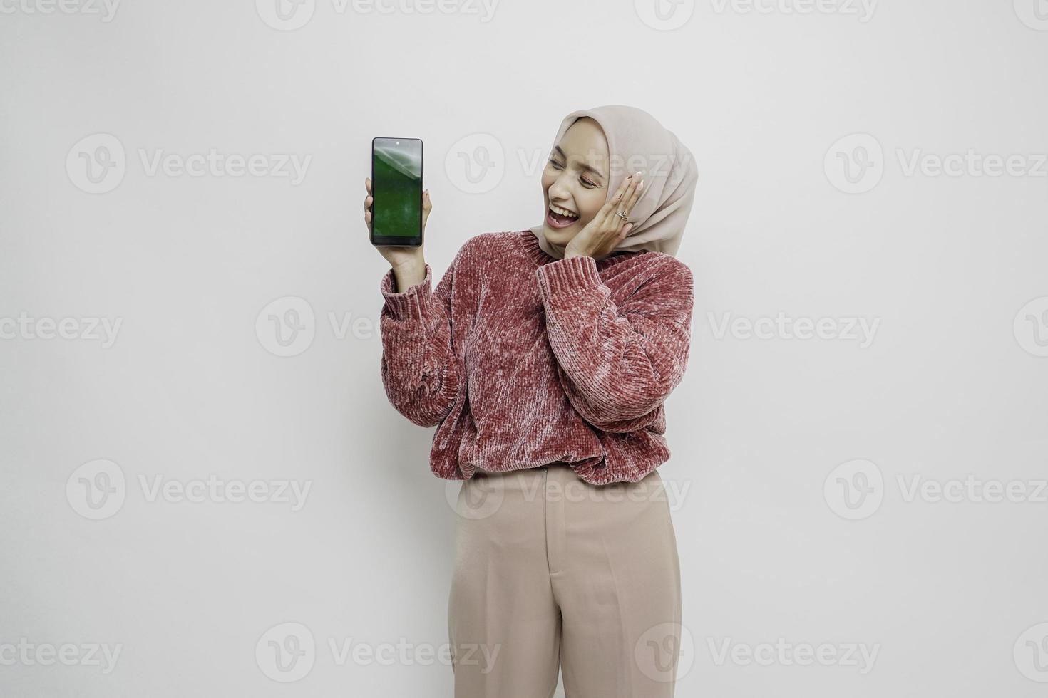 Excited Asian Muslim woman wearing pink sweater and hijab pointing at the copy space beside her while holding her phone, isolated by white background photo