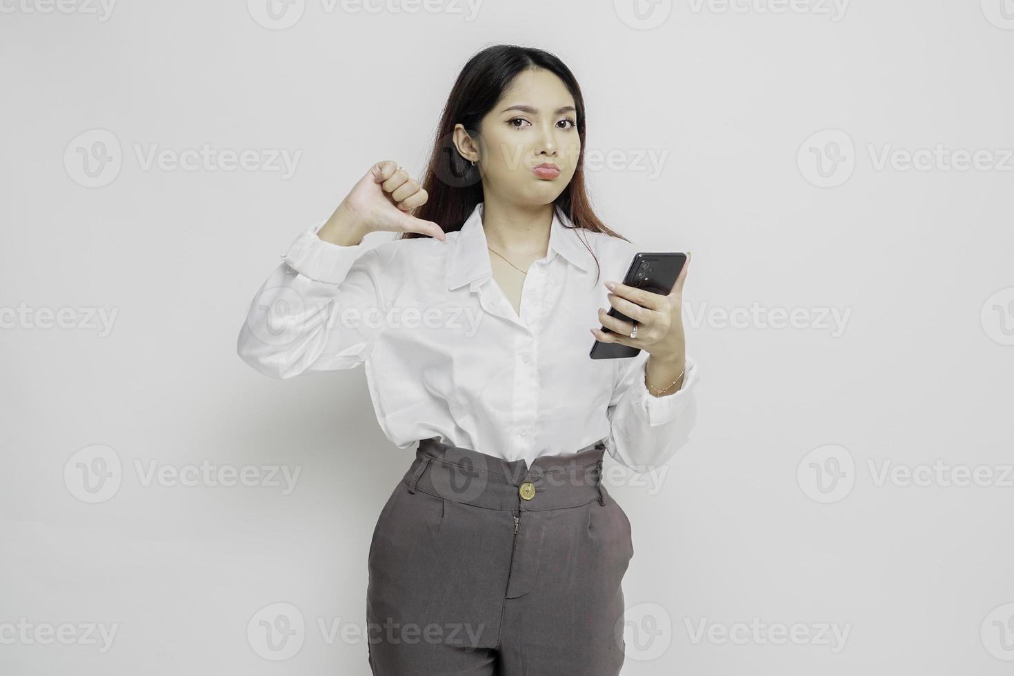 una mujer asiática decepcionada con pantalones blanco da un gesto de desaprobación con la mano hacia abajo, aislada de fondo blanco foto