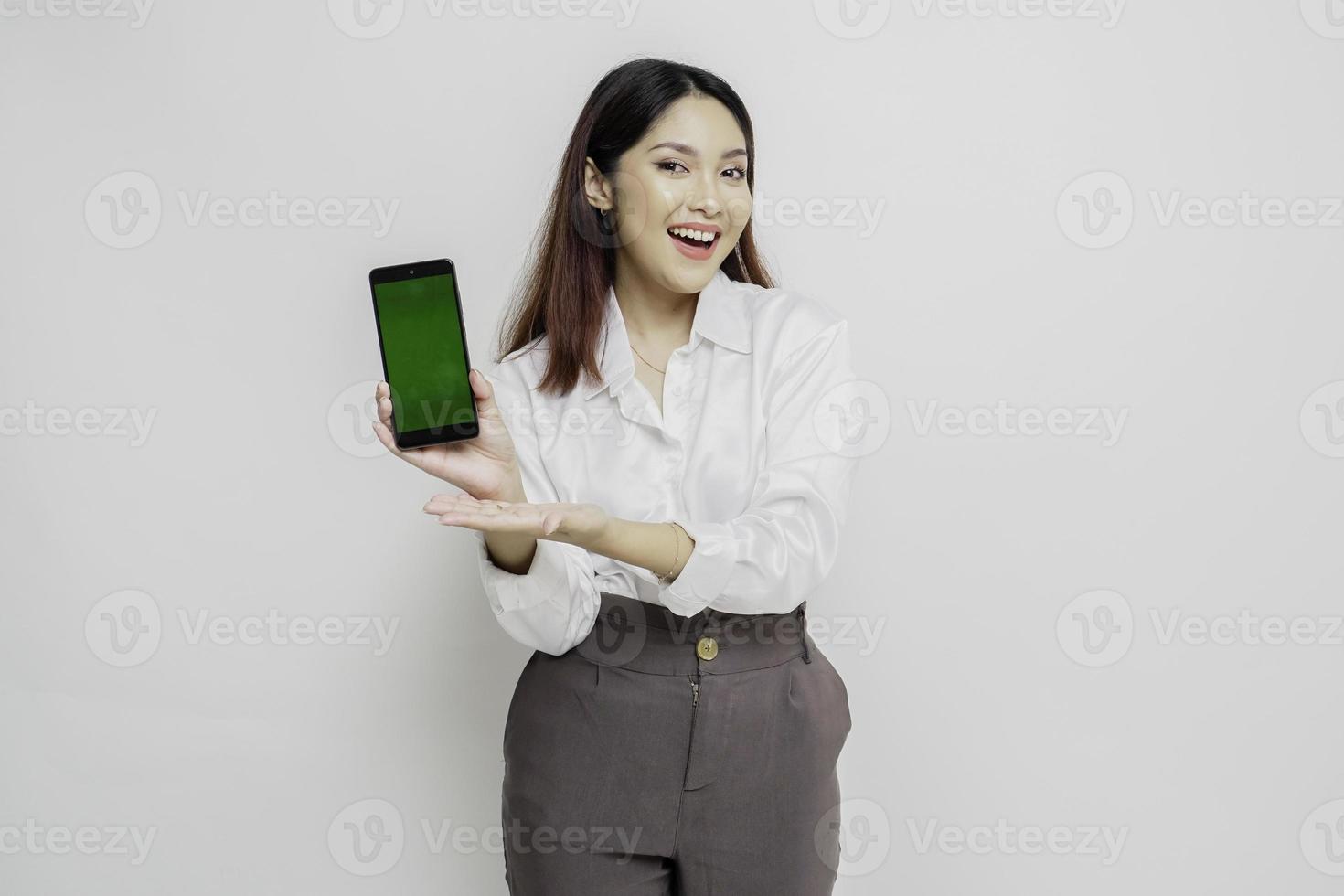 mujer asiática emocionada con pantalones blanco apuntando al espacio de copia a su lado mientras sostiene su teléfono, aislada por fondo blanco foto