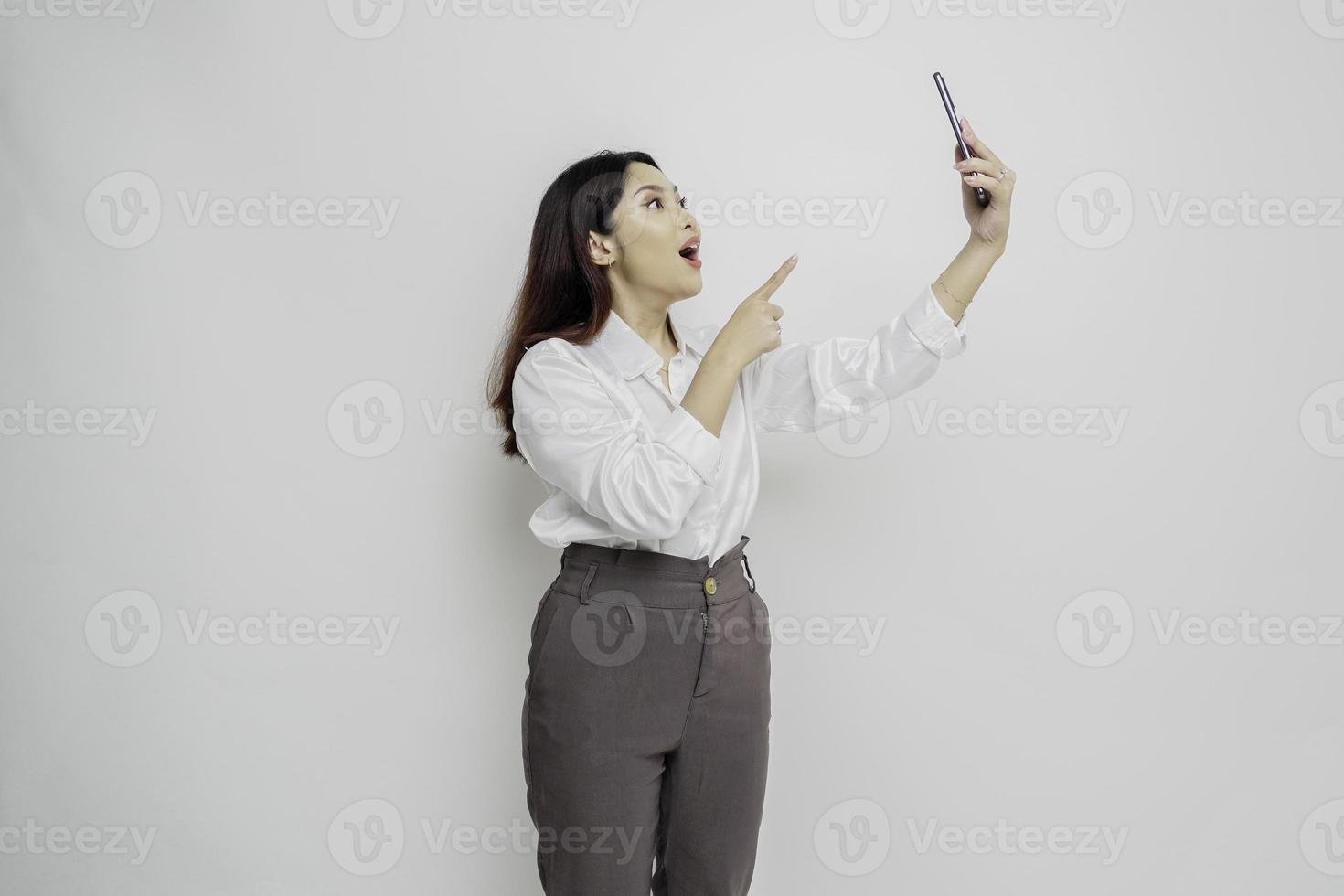 Shocked Asian woman wearing white shirt and holding her phone, isolated by white background photo